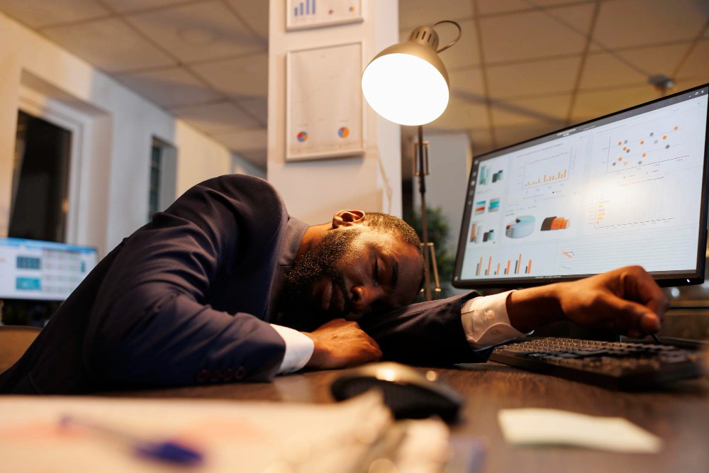 A professional man in a suit asleep at his desk with charts displayed on dual monitors, symbolising burnout and workplace fatigue.