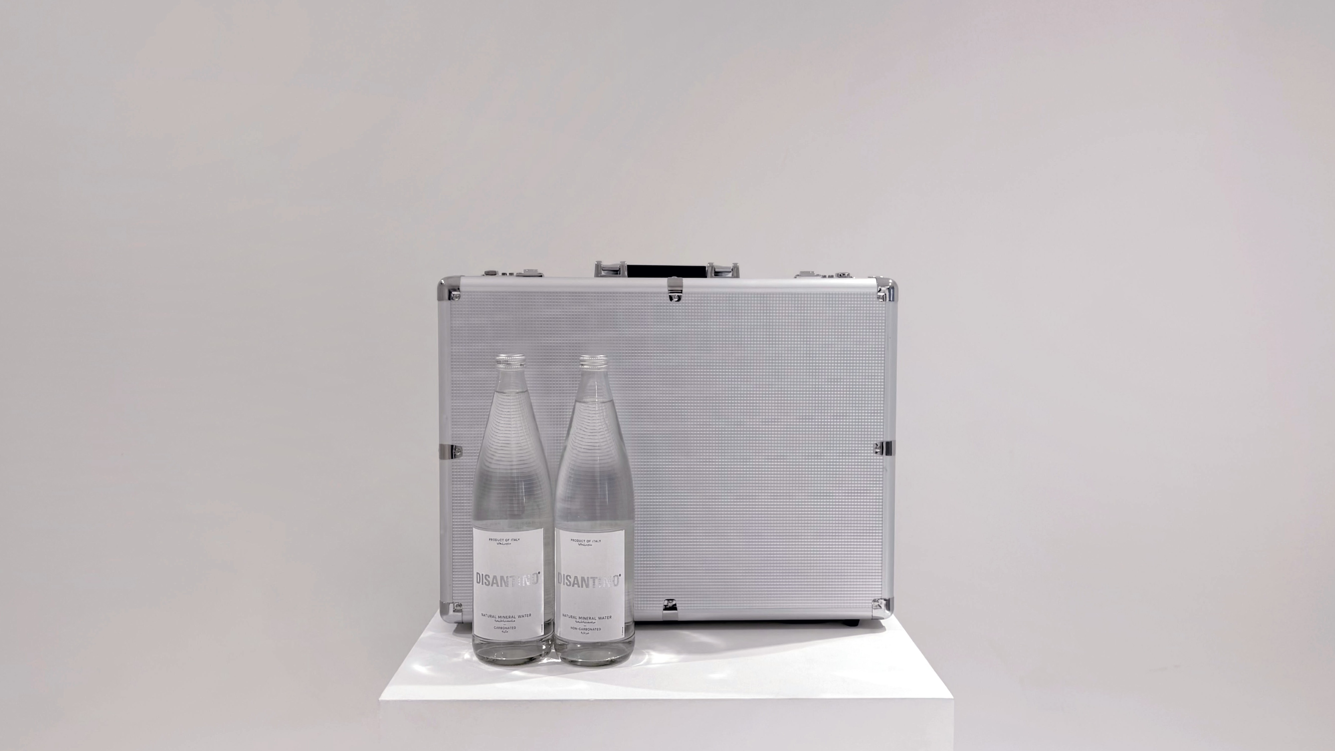 Two clear glass bottles of DISANTINO water stand in front of a large, textured silver metal briefcase. Both bottles and the briefcase are resting on a white square pedestal against a plain light grey background.