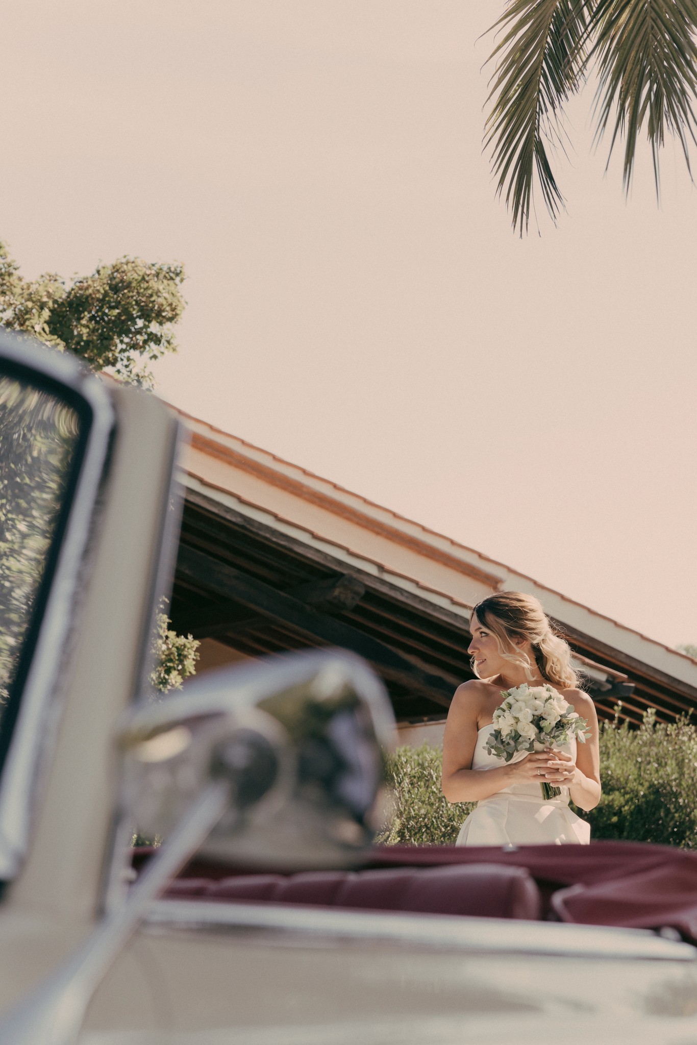Sposa in abito bianco con bouquet tra i filari di Bolgheri, vista dal finestrino della Mercedes d'epoca al tramonto