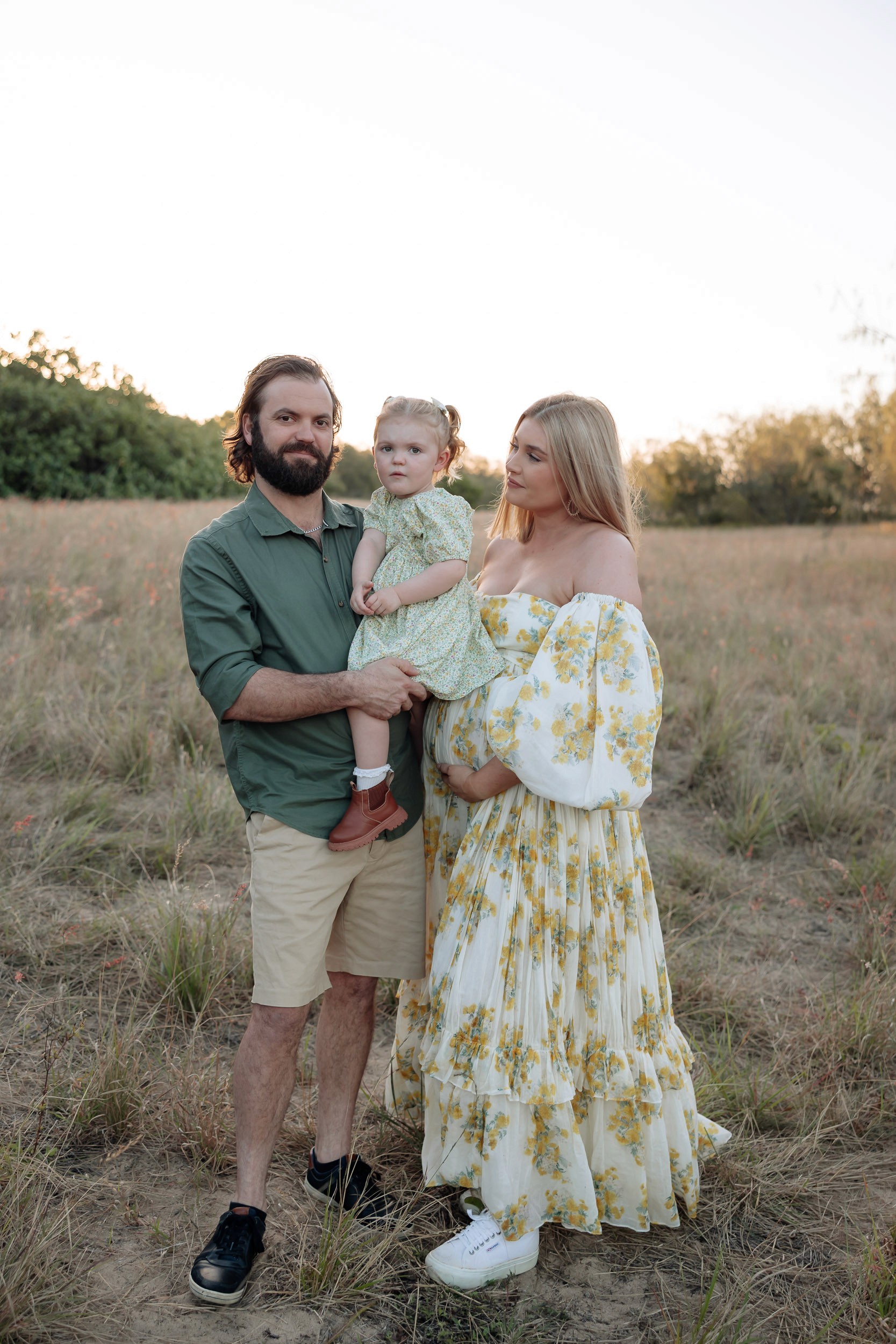 Pregnant mother standing in long grass at sunset with husband and young child during maternity photoshoot in Mackay