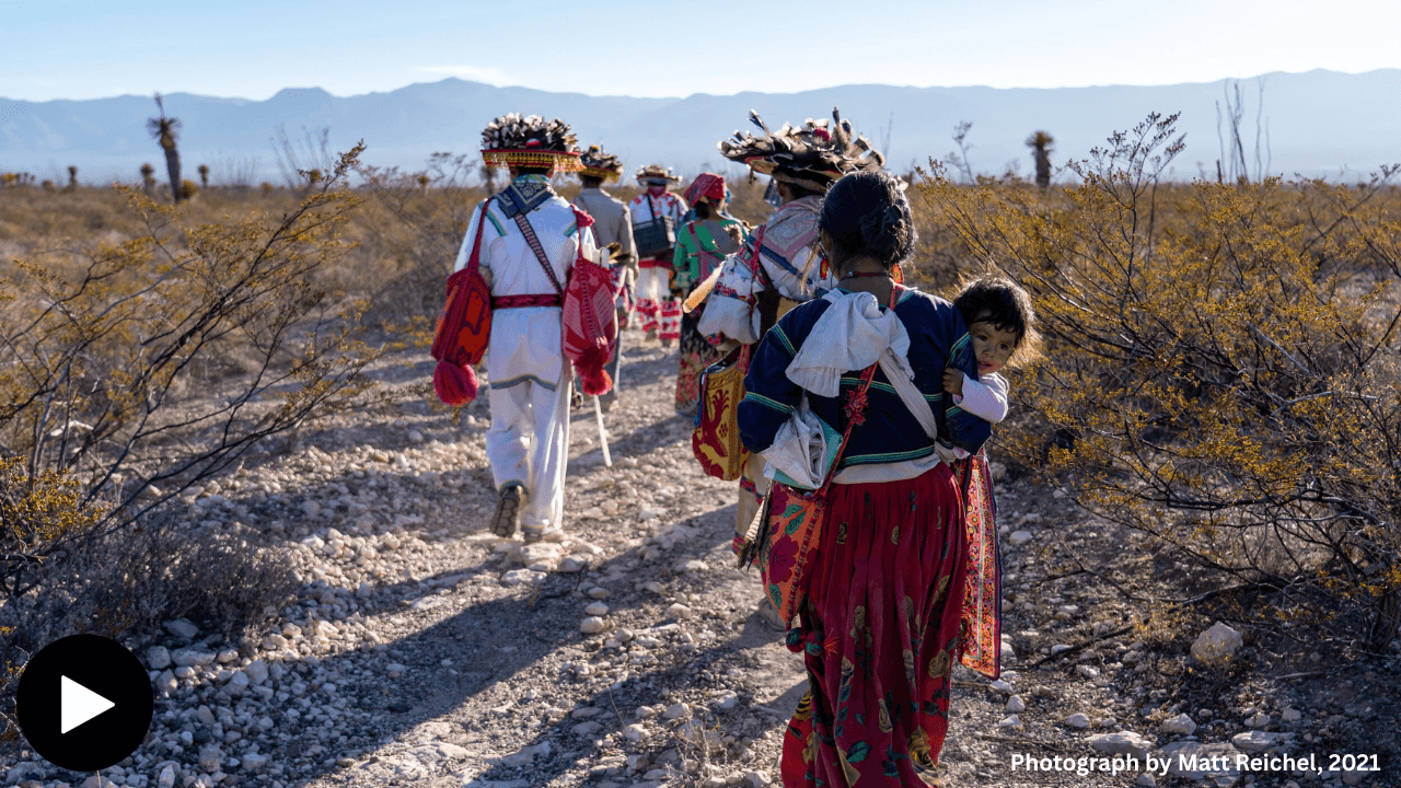 Image shows a photogragh of a group of Wixárika people travelling along a path in the desert. The two figures closest to the viewer is a mother carrying her daughter.