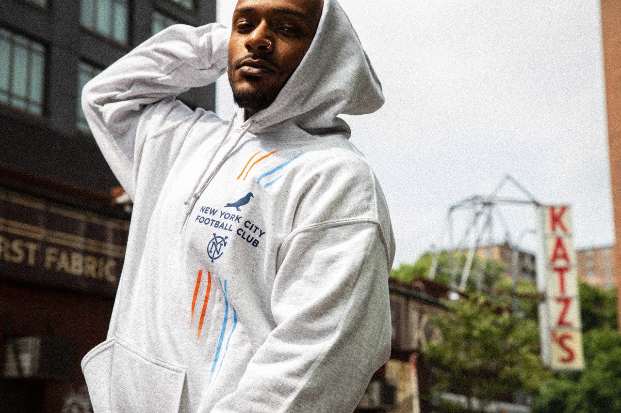 Model in a light gray hoodie from the Staple x NYCFC collaboration, photographed on a rooftop with the city in the background.