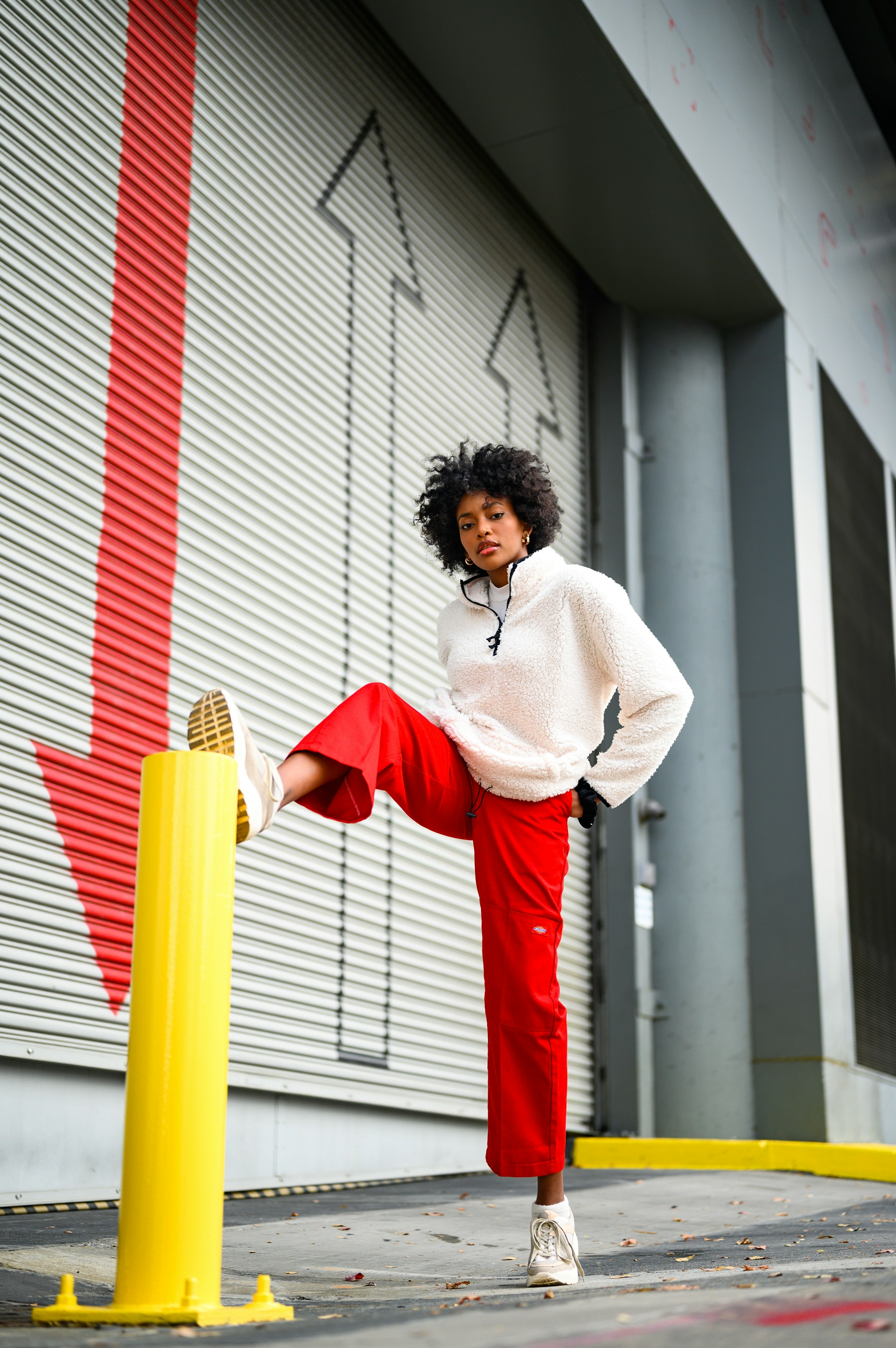 An action shot of the woman wearing a white fleece and red pants, resting her foot on a bright yellow bollard. She is posed in front of a grey warehouse door with large red and white arrow graphics.