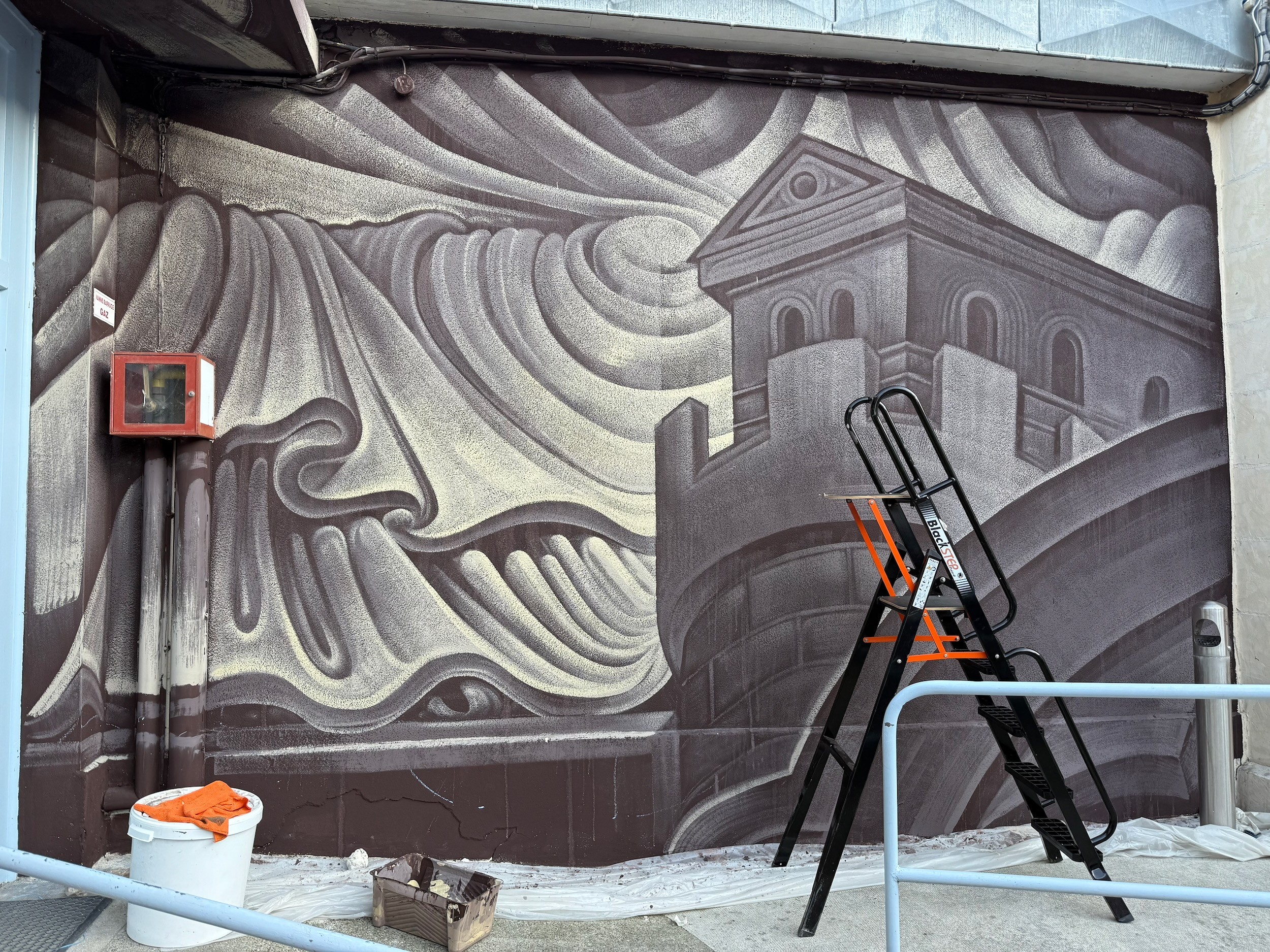 Folds and a carved tower in grey and plum fill a wall, a stepladder and buckets at its foot.