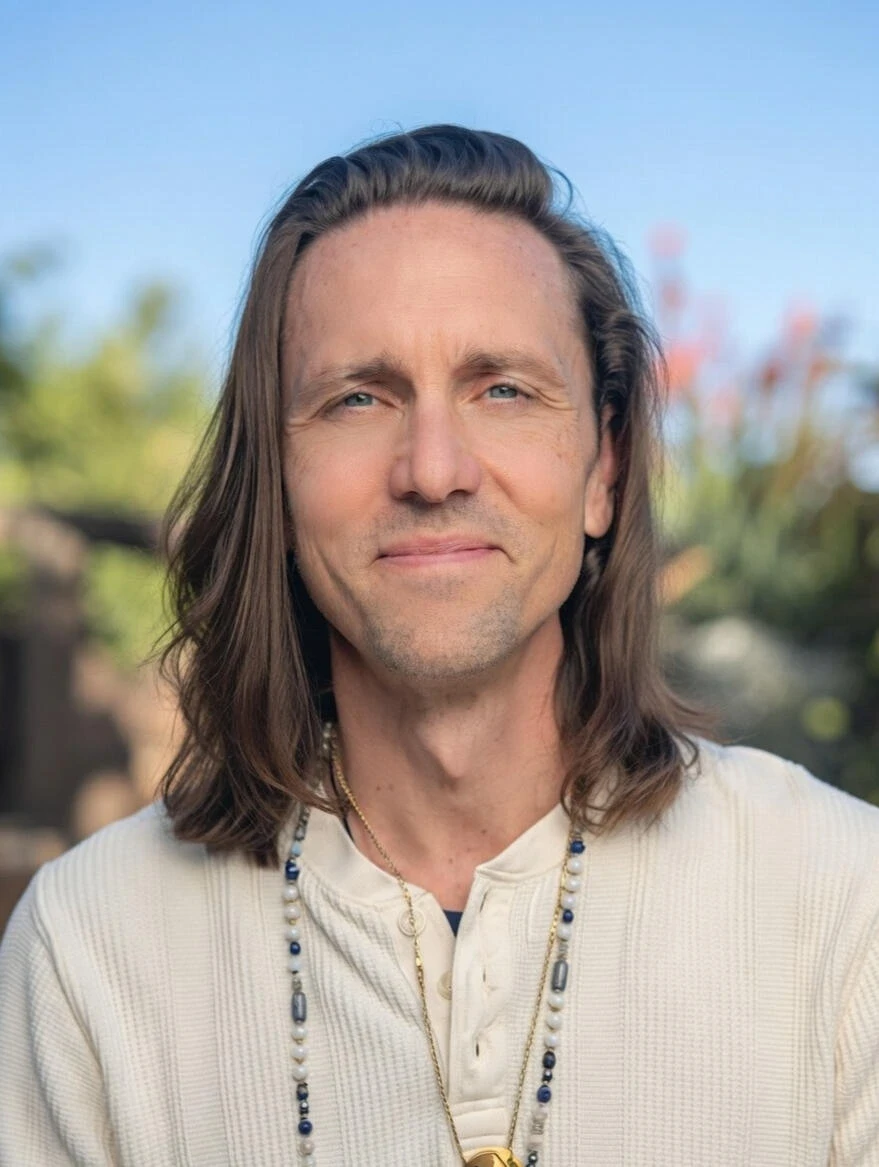 Portrait of sean with shoulder-length brown hair, wearing a cream textured shirt and beaded necklace, softly smiling against a blurred outdoor background with greenery and sky.