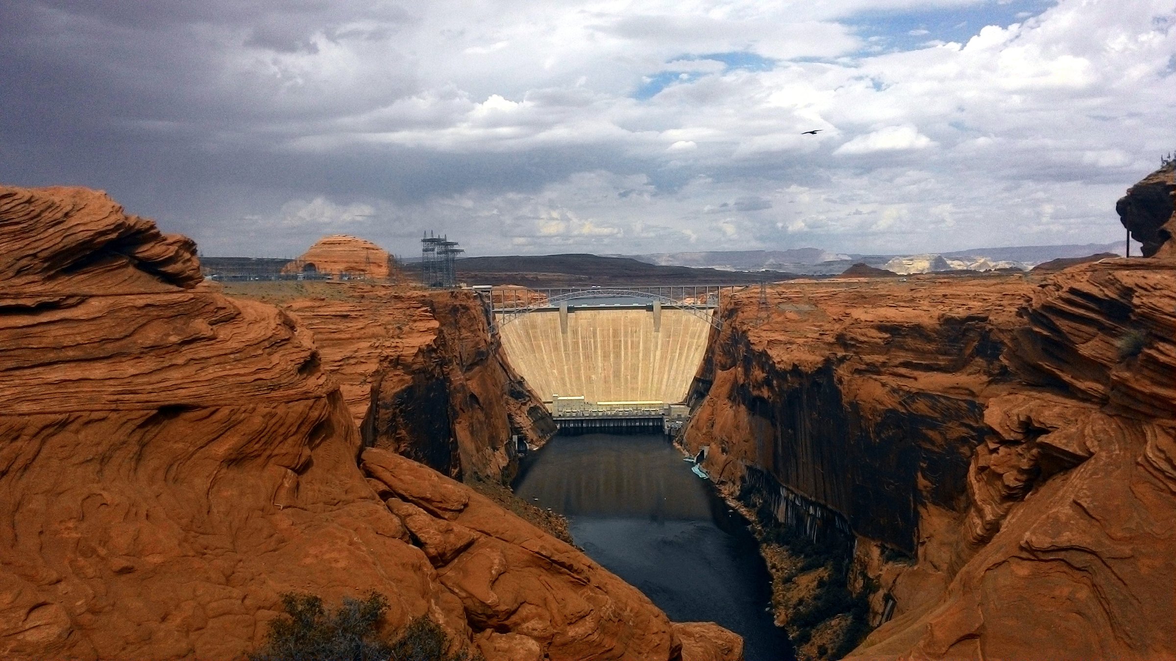 Glen Canyon Dam seen from a sandstone overlook downstream, the pale curved dam face wedged between red canyon walls, the Colorado River dark and still below it under a cloudy sky.