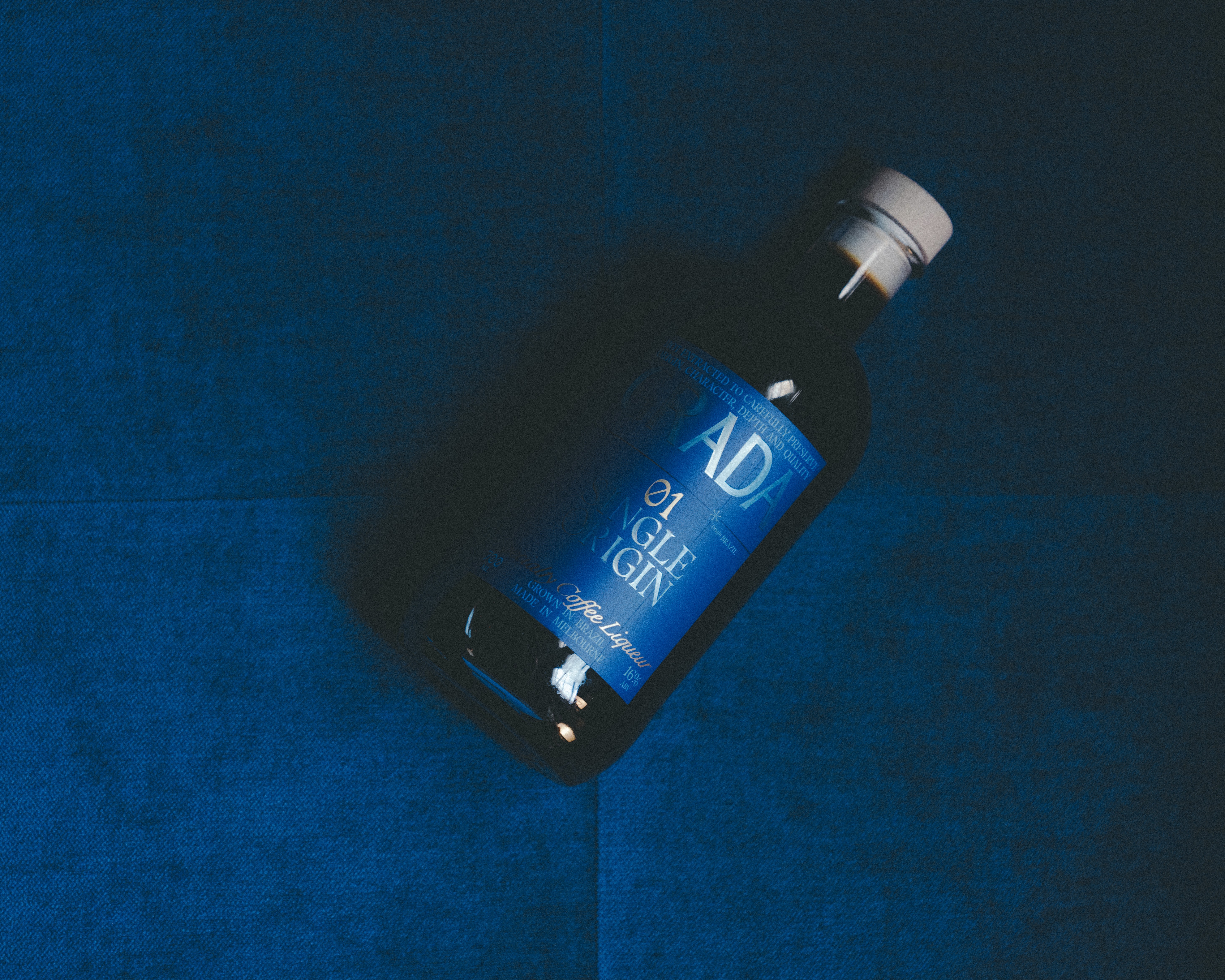 A bottle with a blue label featuring the words "Grade 01 Single Origin" lies on a plush, dark blue circular surface, creating a sophisticated and luxurious aesthetic.