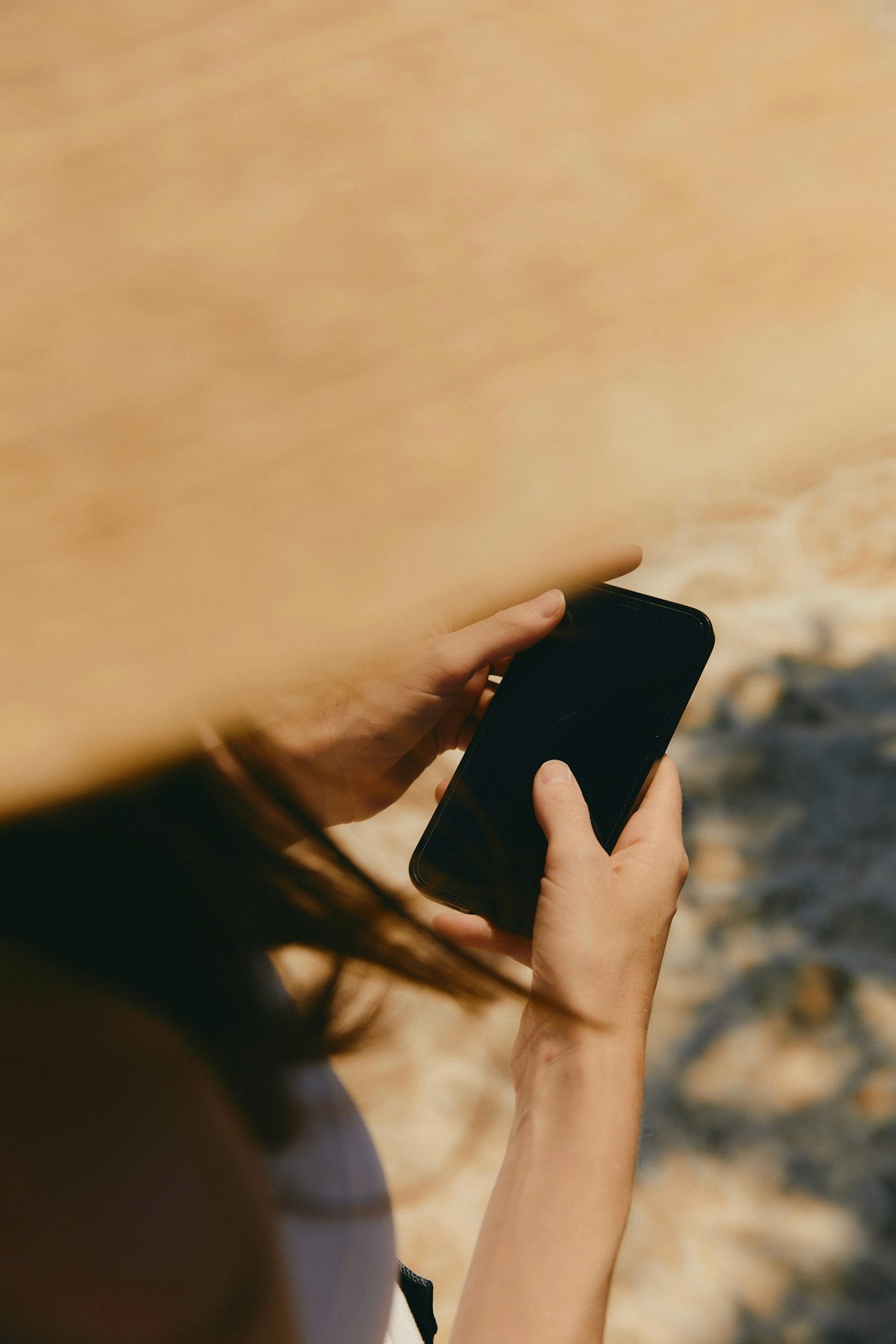Des mains tiennent un smartphone noir à l'écran éteint, sur un fond brun clair flou avec des taches de soleil.