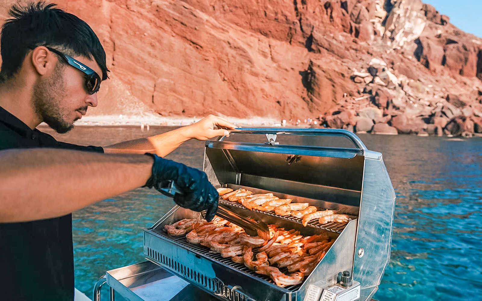Grilling shrimp on a catamaran with Santorini's red cliffs in the background.