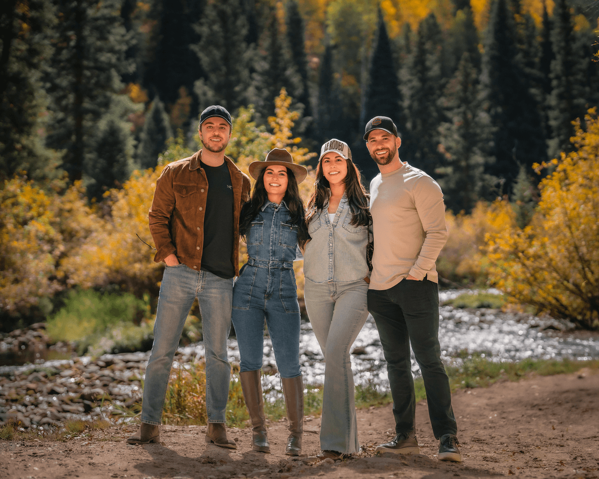 Four friends in hats posing beside a stream surrounded by golden autumn foliage