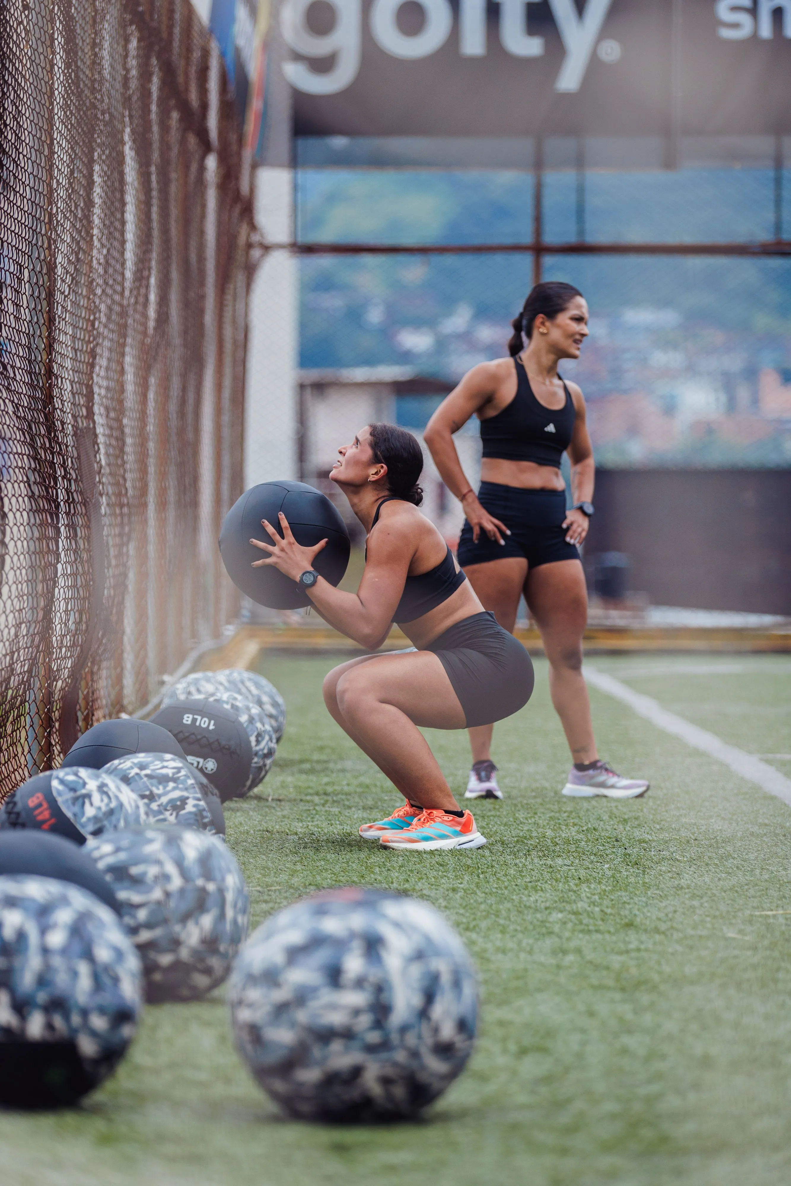 Two women exercise outdoors on a sports field, with one performing squats using a medicine ball while the other observes, surrounded by scattered patterned medicine balls; keywords: outdoor exercise, medicine ball workout.