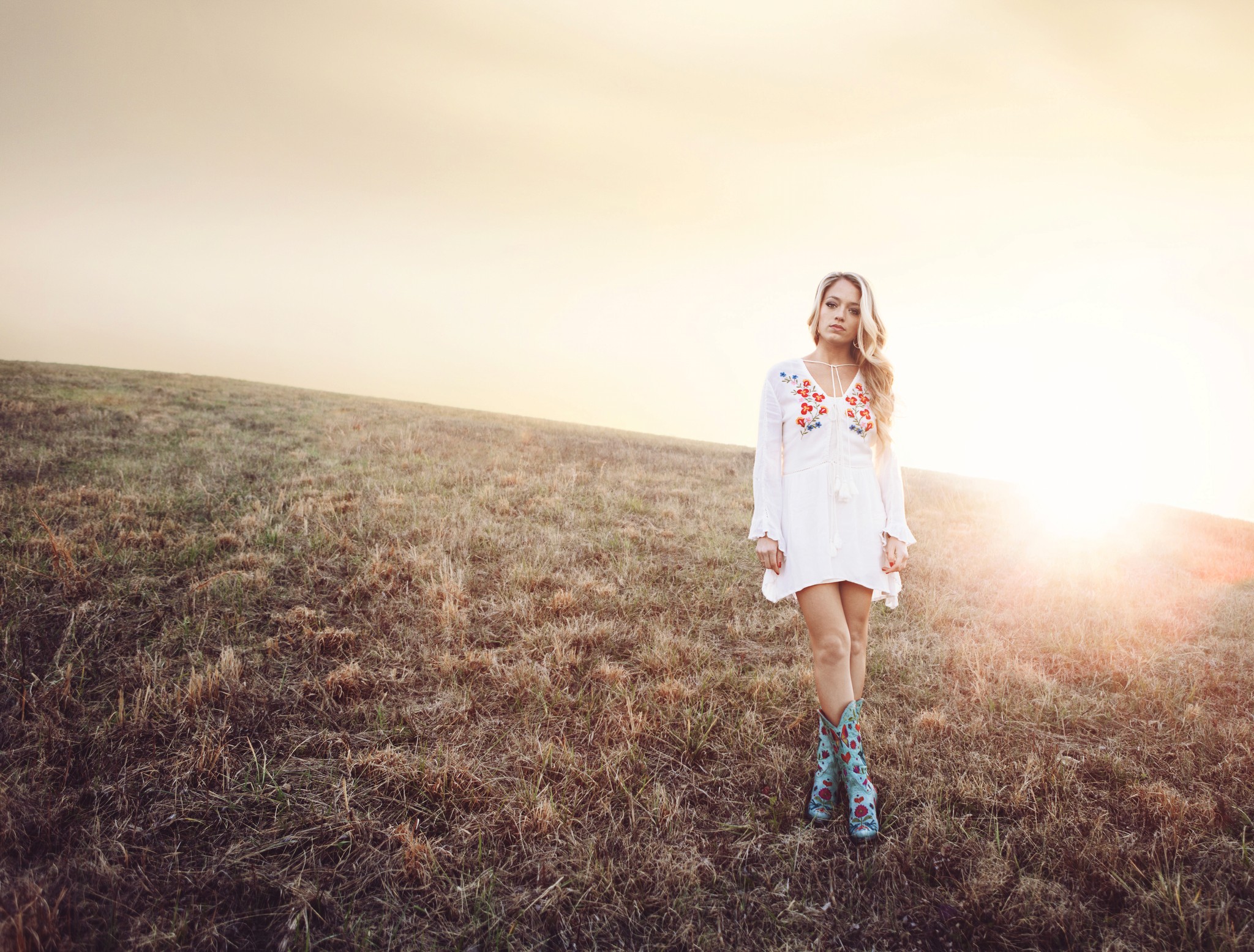 Editorial portrait of country music artist Jordyn Mallory standing in an open field at golden hour, wearing a white embroidered dress and boots.