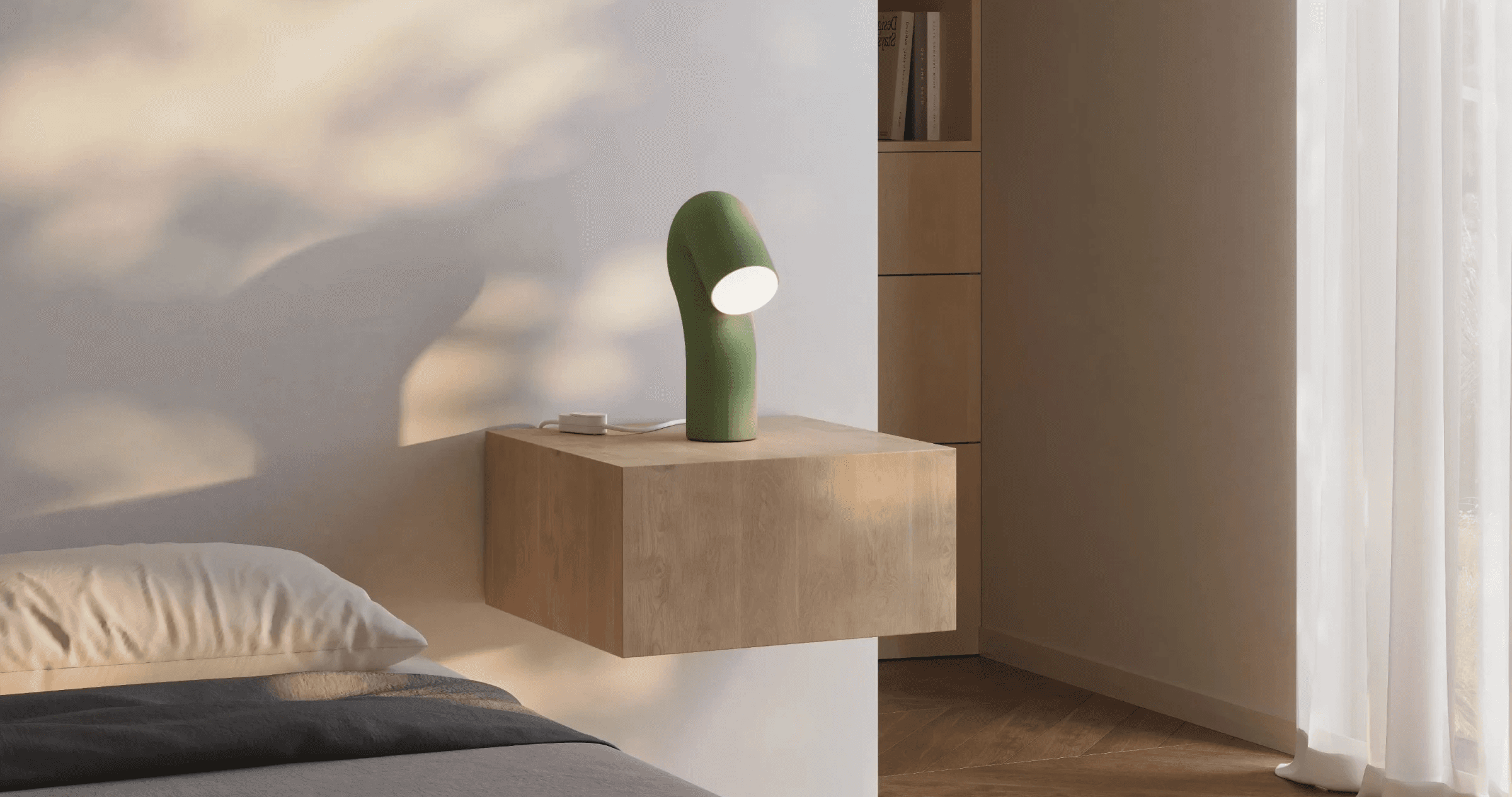 Green bent-tube table lamp glowing on a floating wood nightstand beside a bed, with leaf shadows on the wall