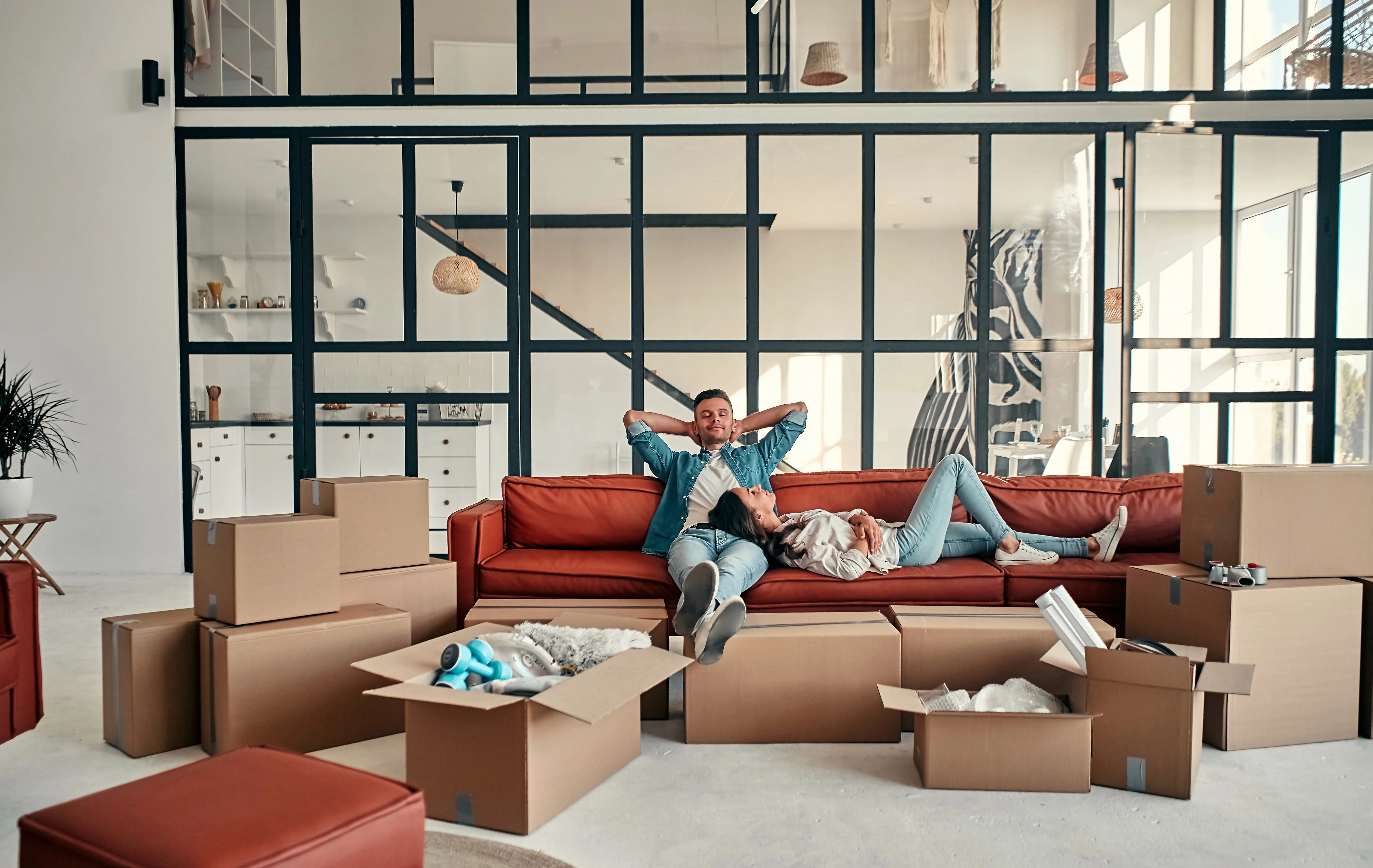 A couple laying on a red couch, taking a break from unpacking moving boxes.