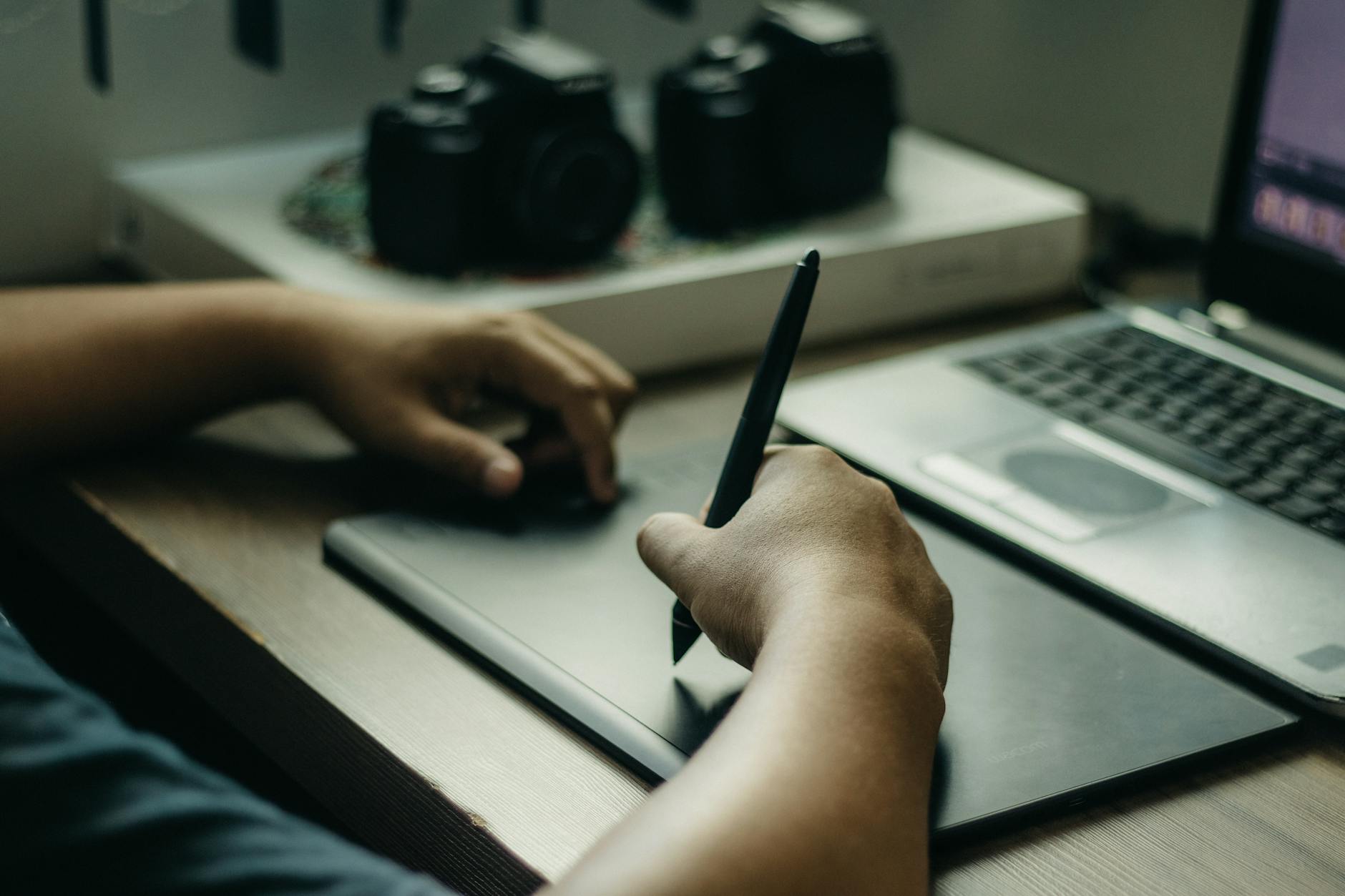 A close-up of a student's hands typing on a laptop next to an open textbook and a cup of pens on a wooden desk.