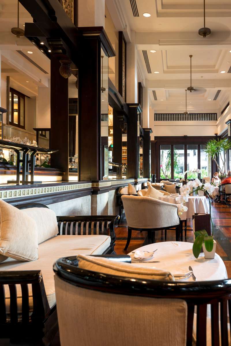 A high-ceilinged restaurant interior with dark wood beams, white tablecloths, and a long bar counter under warm lighting.