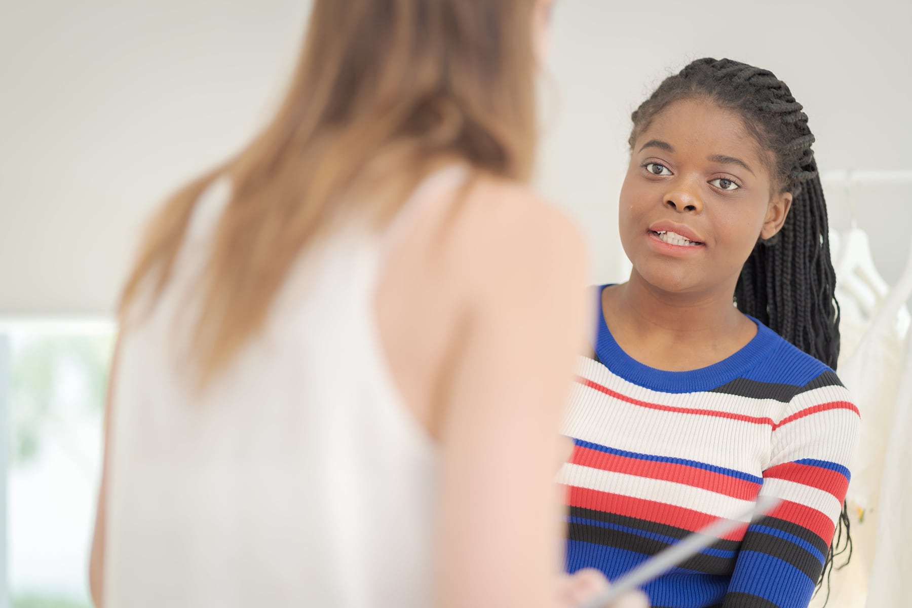 A woman with braided hair speaks to another person in a bright room, appearing engaged in a discussion. Clothing racks are visible in the background.