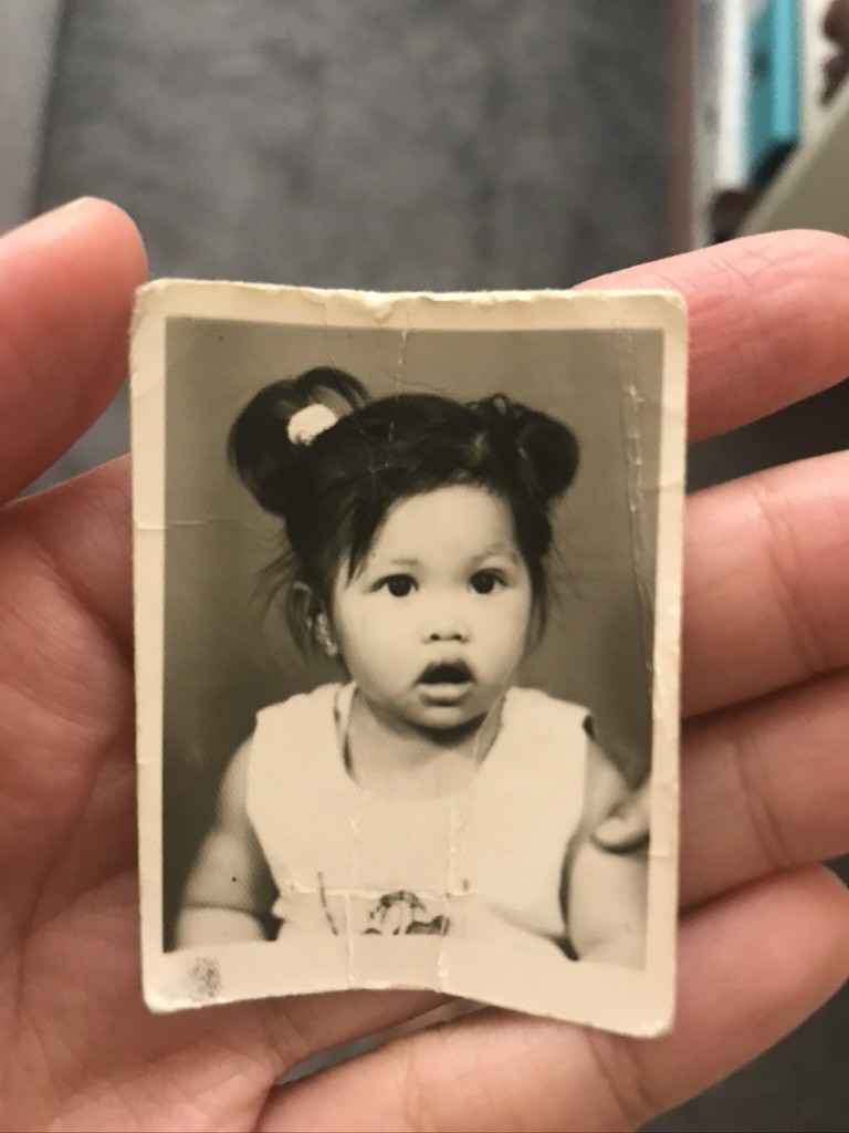 Una persona con una camiseta negra y pantalones de color claro está de pie en la cima rocosa de una montaña al atardecer, sosteniendo una cámara frente a su cara para tomar una foto. Un mar de nubes se extiende detrás de ellos, iluminado por la luz dorada del sol poniente.