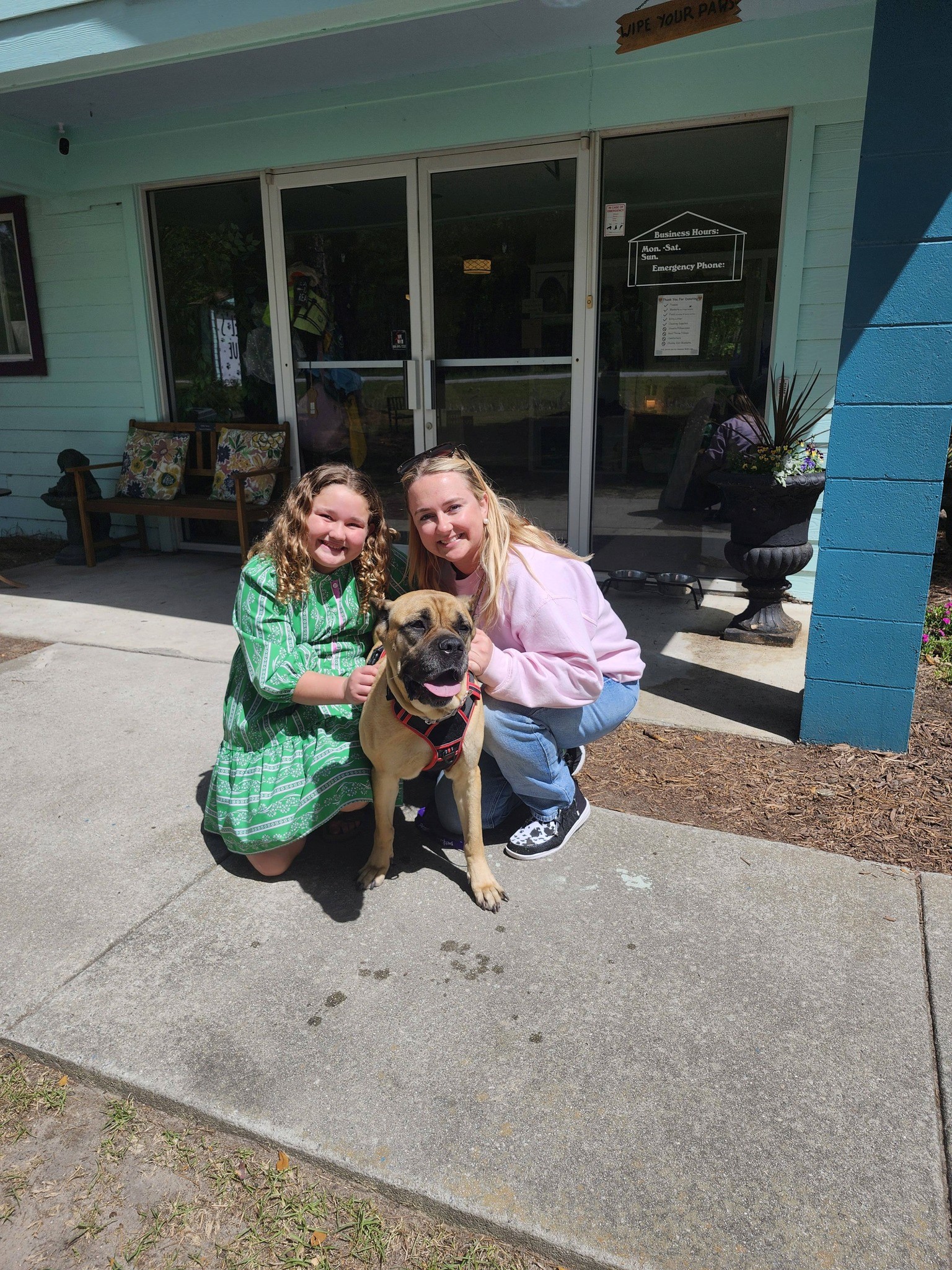 Two people crouch outside a building, smiling while holding and petting a small dog.