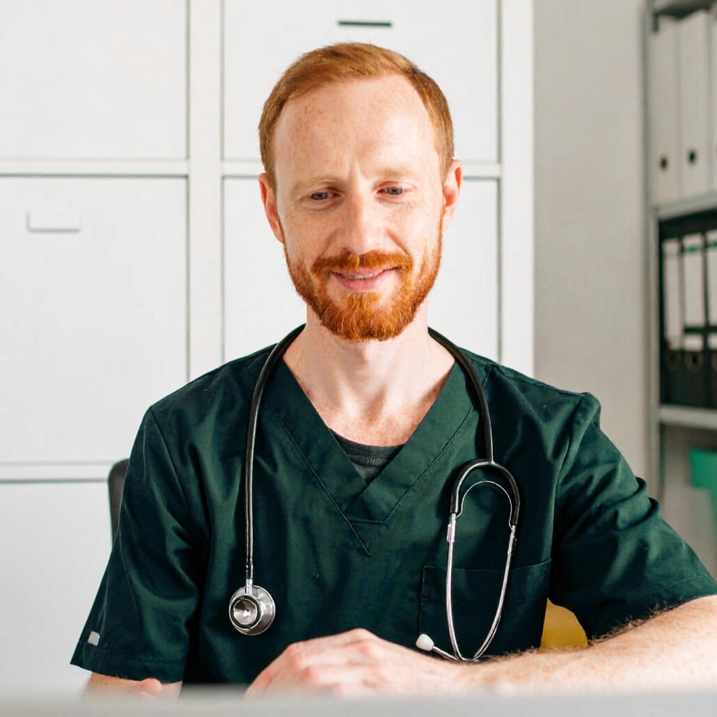 Red-haired man in green scrubs with stethoscope smiles gently in an office setting with cabinets and medical