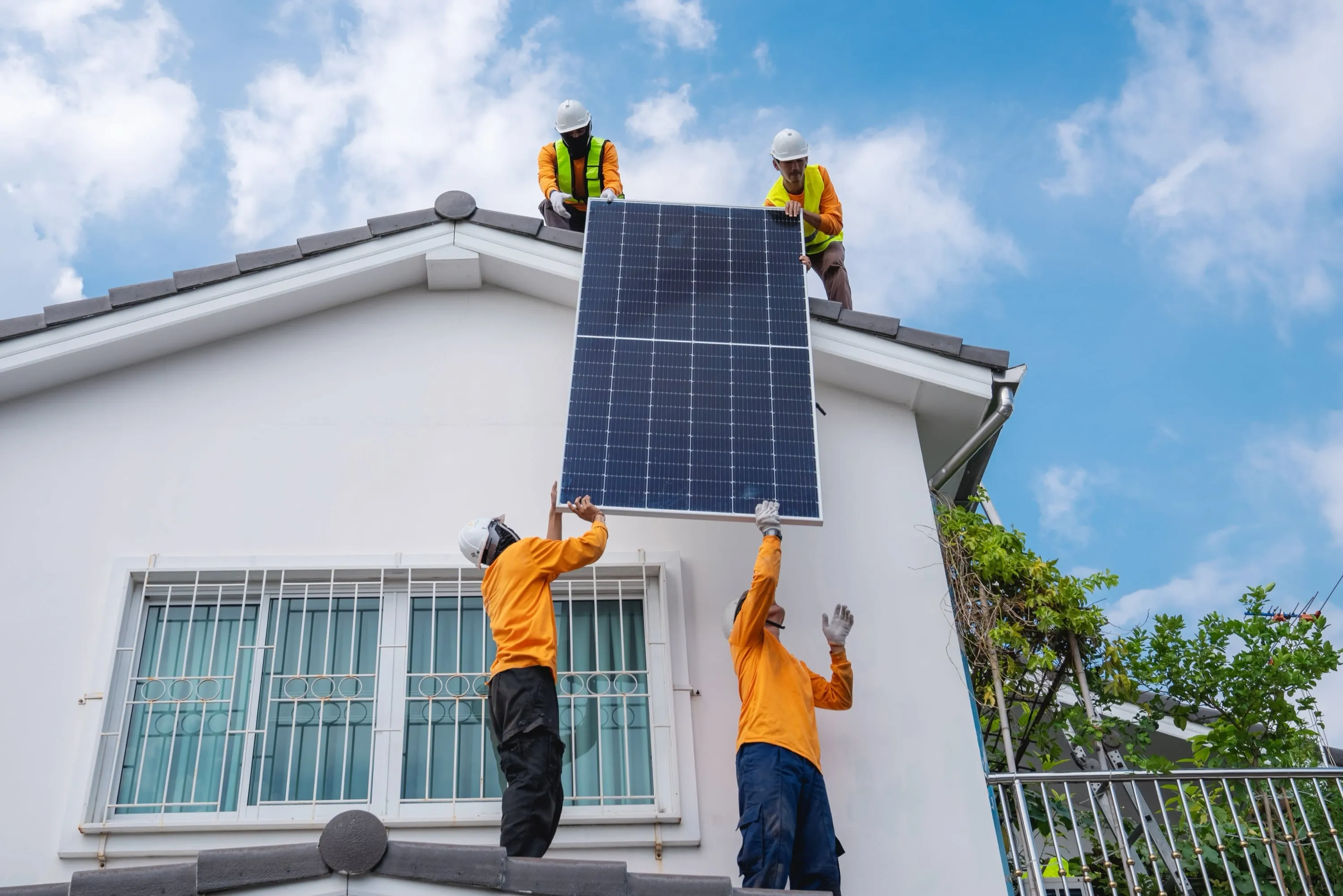 Vier Arbeiter in Sicherheitsausrüstung und orangefarbenen Hemden installieren ein großes Solarpanel auf dem Dach eines weißen Hauses vor strahlend blauem Himmel und machen damit auf nachhaltige Energie und Solartechnologie aufmerksam.