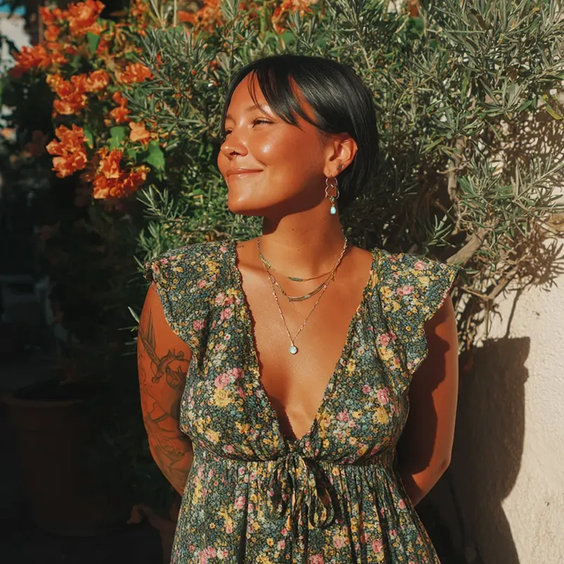 A woman in a floral dress smiles warmly in sunlight, surrounded by lush green foliage and orange flowers. The scene is serene and joyful.