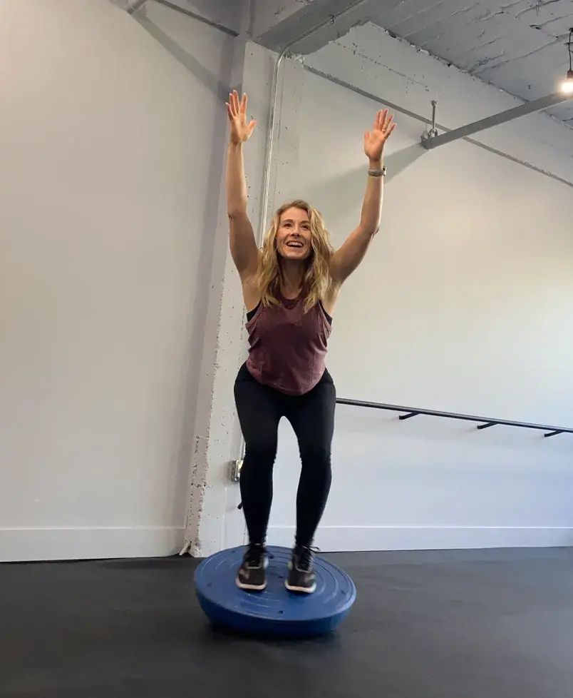 Heather Hausman balances enthusiastically on a Bosu ball, with arms raised, inside a well-lit fitness studio featuring sleek, modern design elements, emphasizing core strength and stability training.