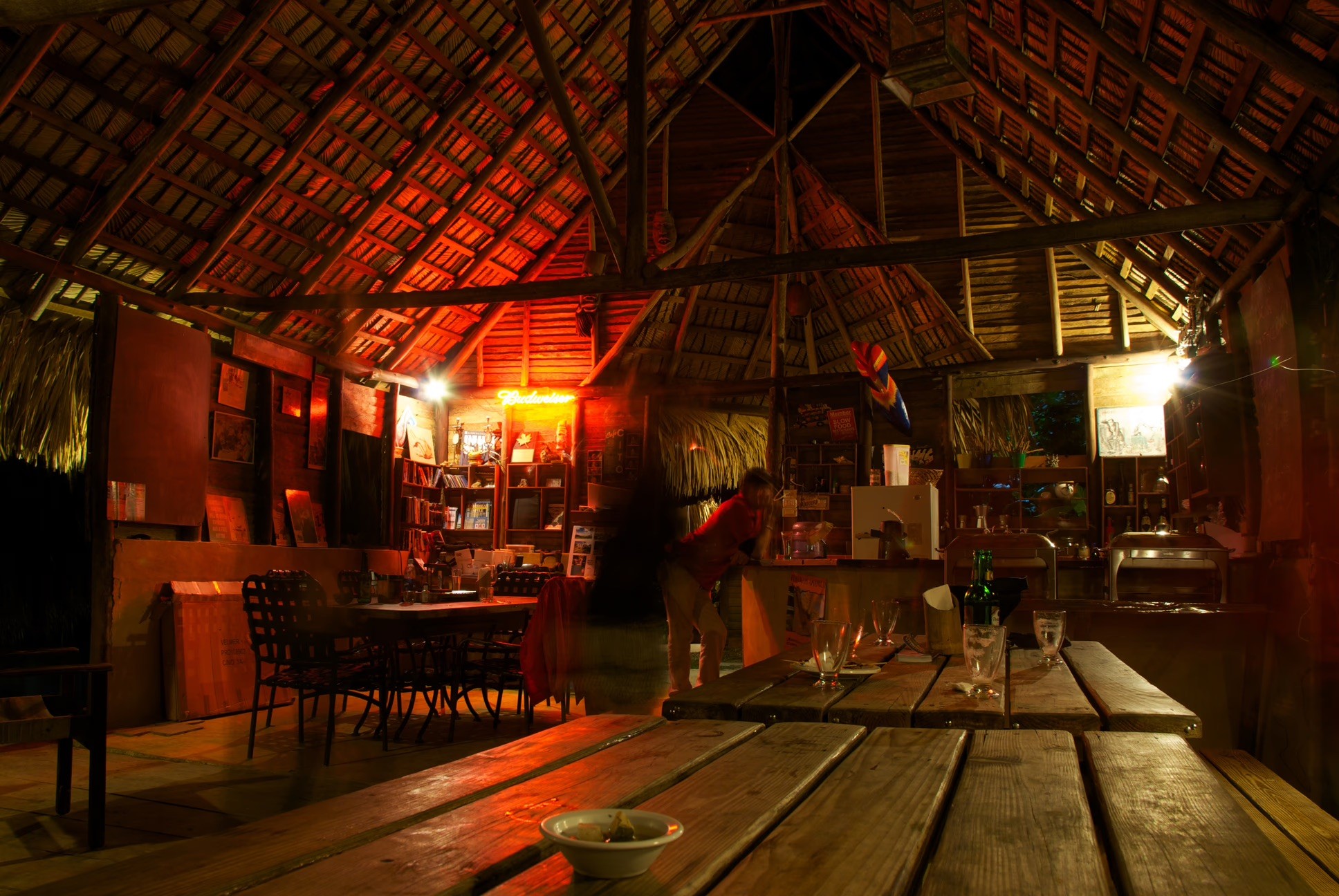 Rustic dining area under a large thatched roof lit at night.