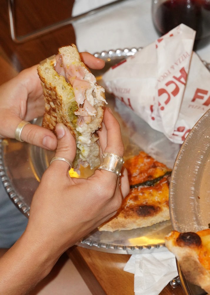 Hand holding a sandwich with pizza slices on a metal tray wearing silver jewelry