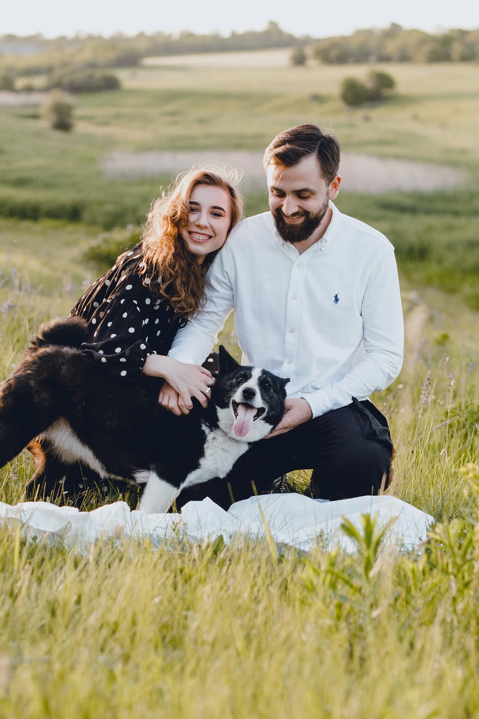 A smiling couple sits on a white blanket in a lush green field.