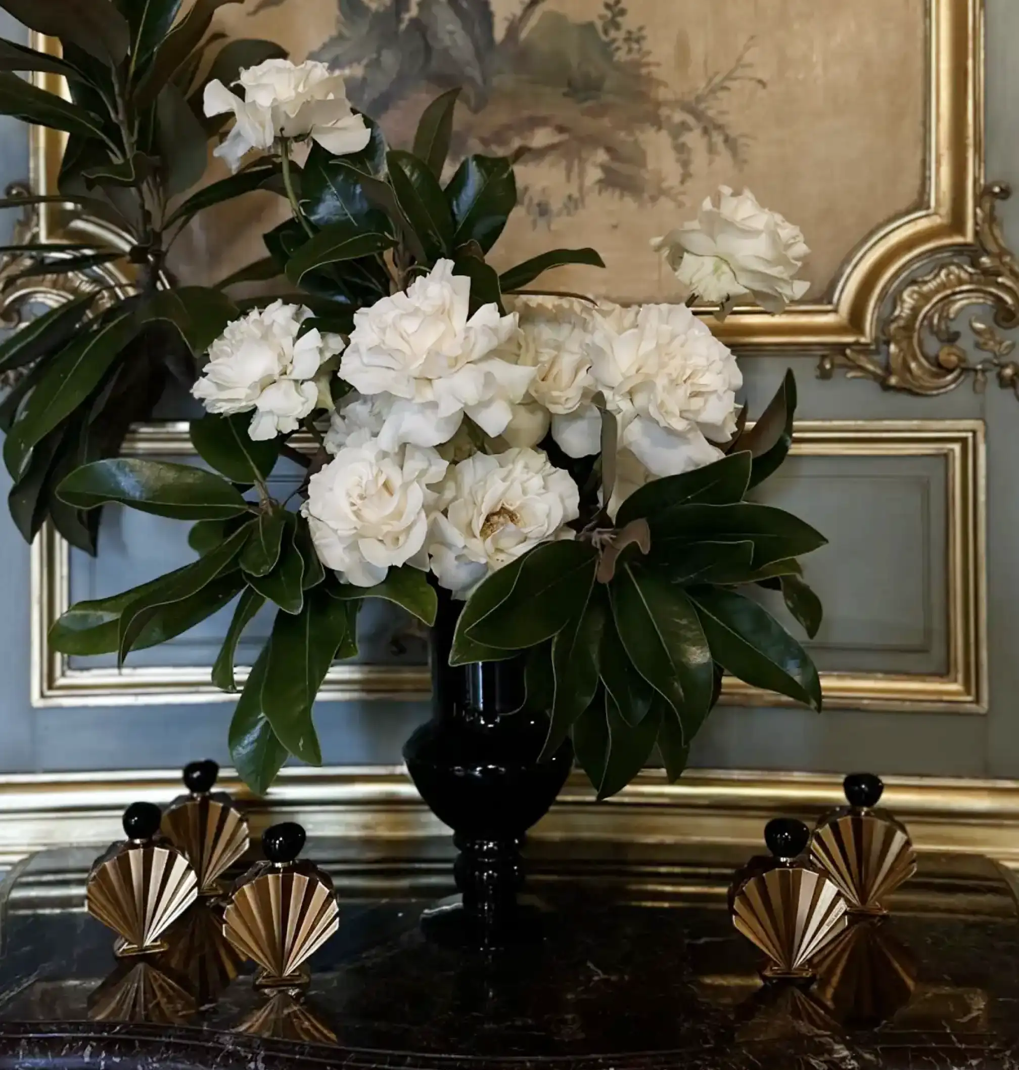 Arreglo floral con flores blancas y hojas verdes brillantes en un jarrón negro, colocado sobre una mesa de mármol oscuro con base dorada tallada, frente a una pared azul con molduras doradas y una pintura clásica enmarcada.