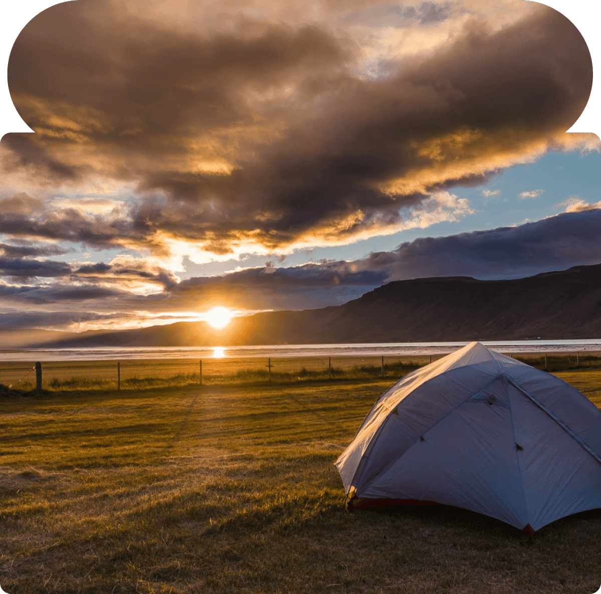 Camping tent on a grassy field at sunset with mountains and dramatic clouds in the background.