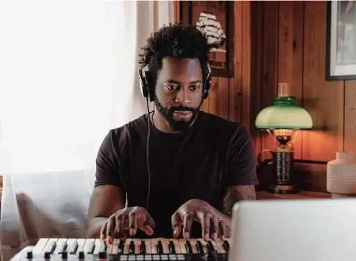 A man with visa approval in a black tshirt making music on a keyboard wearing headphone in a cozy lit room.
