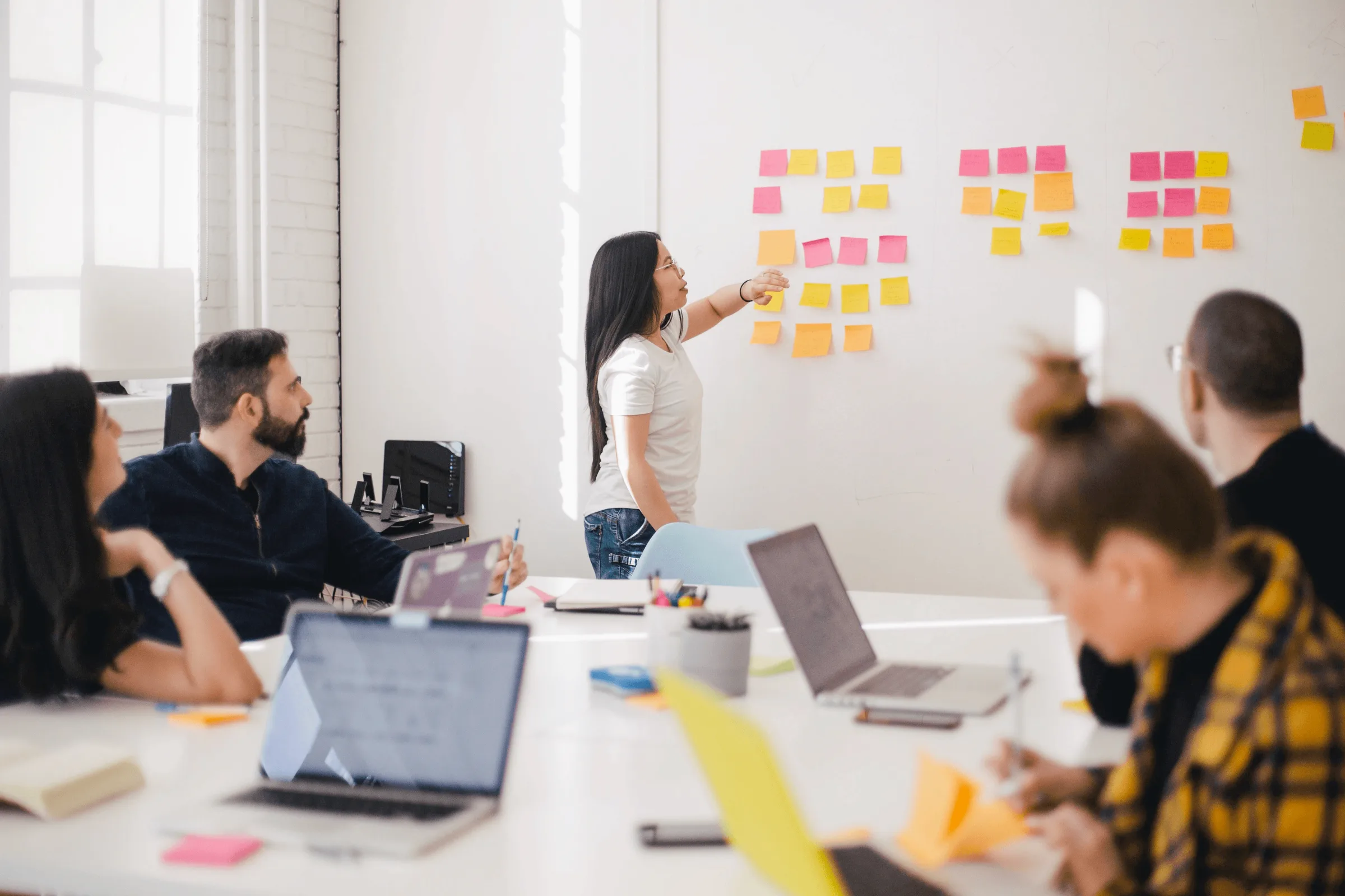 Woman presenting colorful sticky notes on wall to team members seated at conference table with laptops.