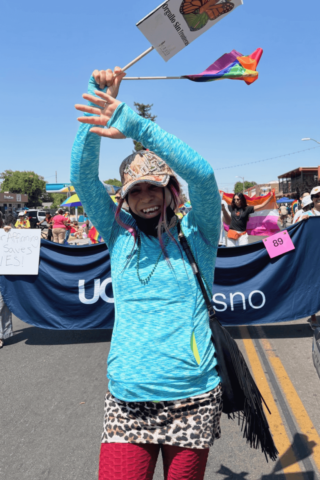 Una persona con ropa vibrante baila alegremente en un desfile animado, sosteniendo una pequeña bandera con un diseño de mariposa, rodeada de pancartas coloridas y personas celebrando bajo un cielo azul despejado.