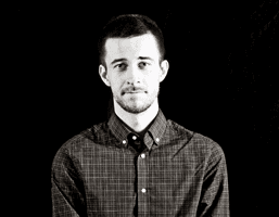 A black and white portrait of a man with short dark hair and a short beard, wearing a dark plaid button-down shirt. The background is solid black.