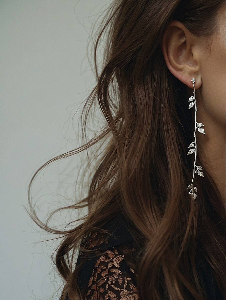 Elegant silver leaf earring on a woman with wavy brown hair and a black patterned top, set against a muted gray background.