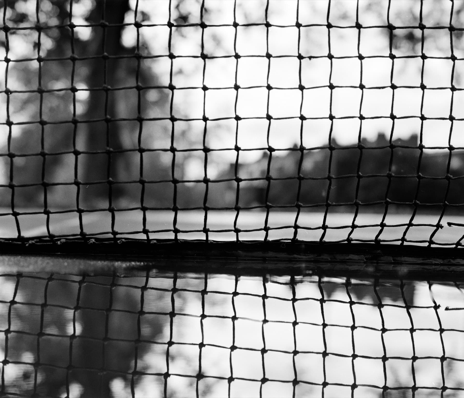 Tennis court net with irregular grid pattern reflected in puddle, mirrored symmetry creating doubled effect, shallow depth of field with bokeh beyond the netting