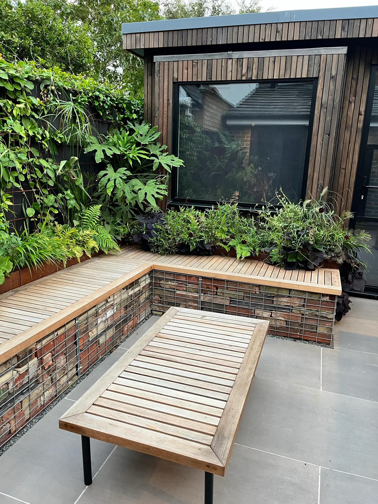 A wooden bench sits on a stone patio near greenery and a building with large windows.