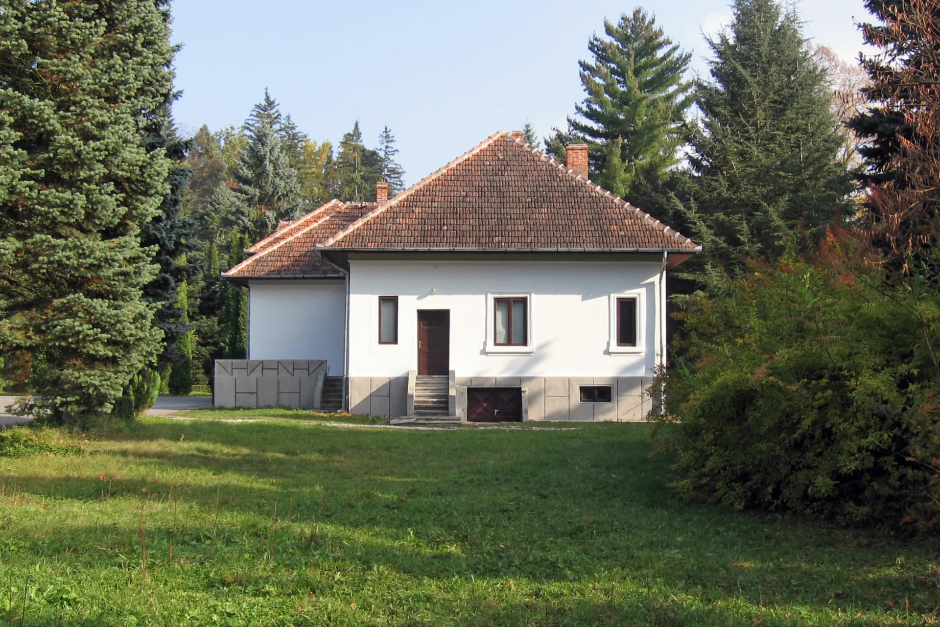 Ein Haus, welches vor seinem Garten fotografiert wurde. Links und rechts sind verschiedenste Bäume und Büsche.