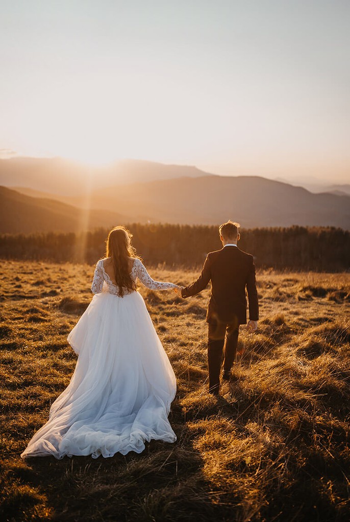 A couple in wedding attire walks hand in hand across a sunlit field.