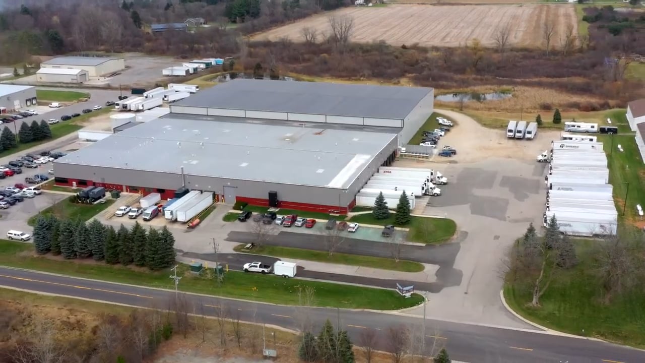 Aerial view of a large industrial warehouse facility surrounded by parked trucks and trailers, with adjacent fields and trees, showcasing a streamlined logistics and distribution hub.