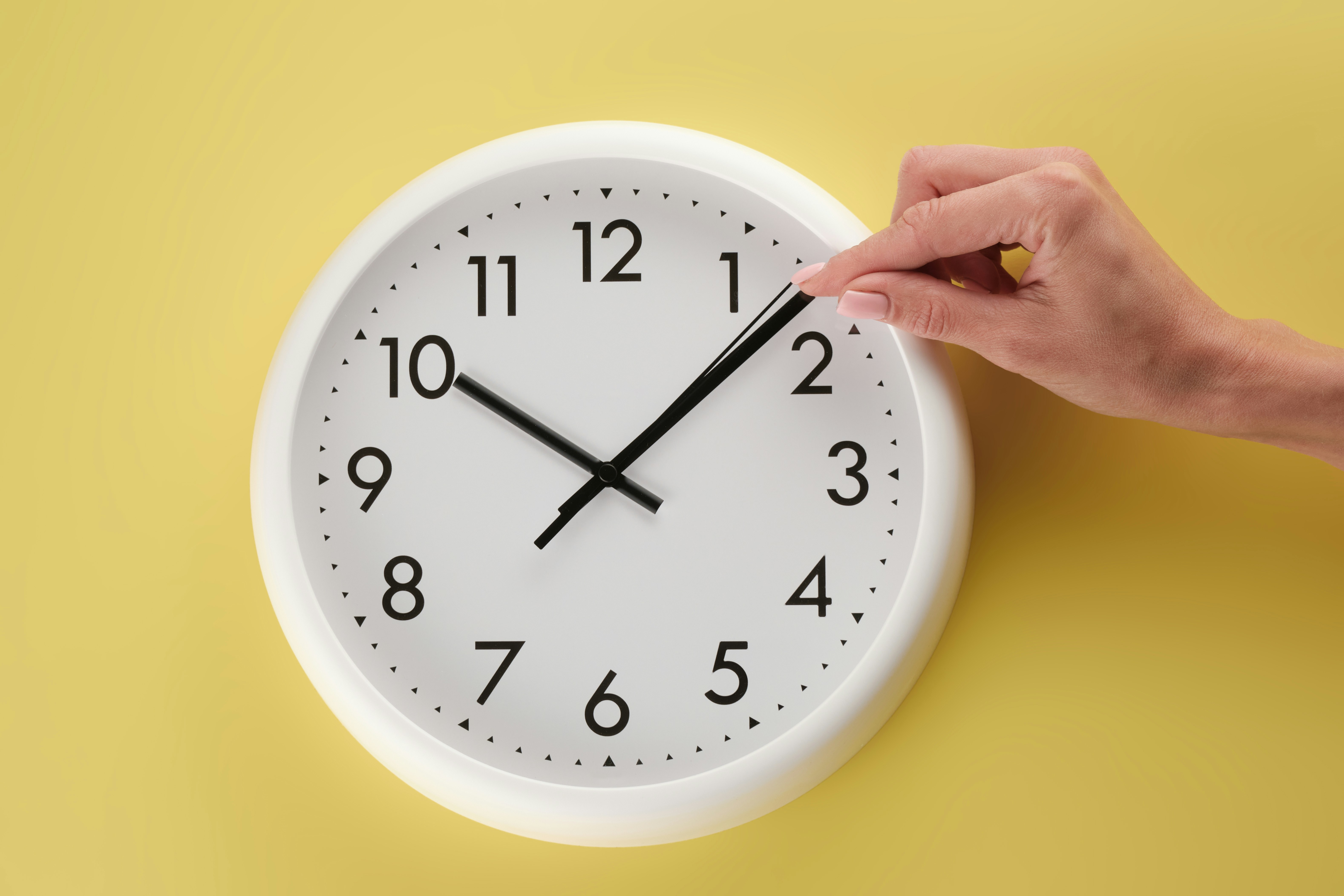 a person's hand adjusting the time on an analog clock against a yellow background