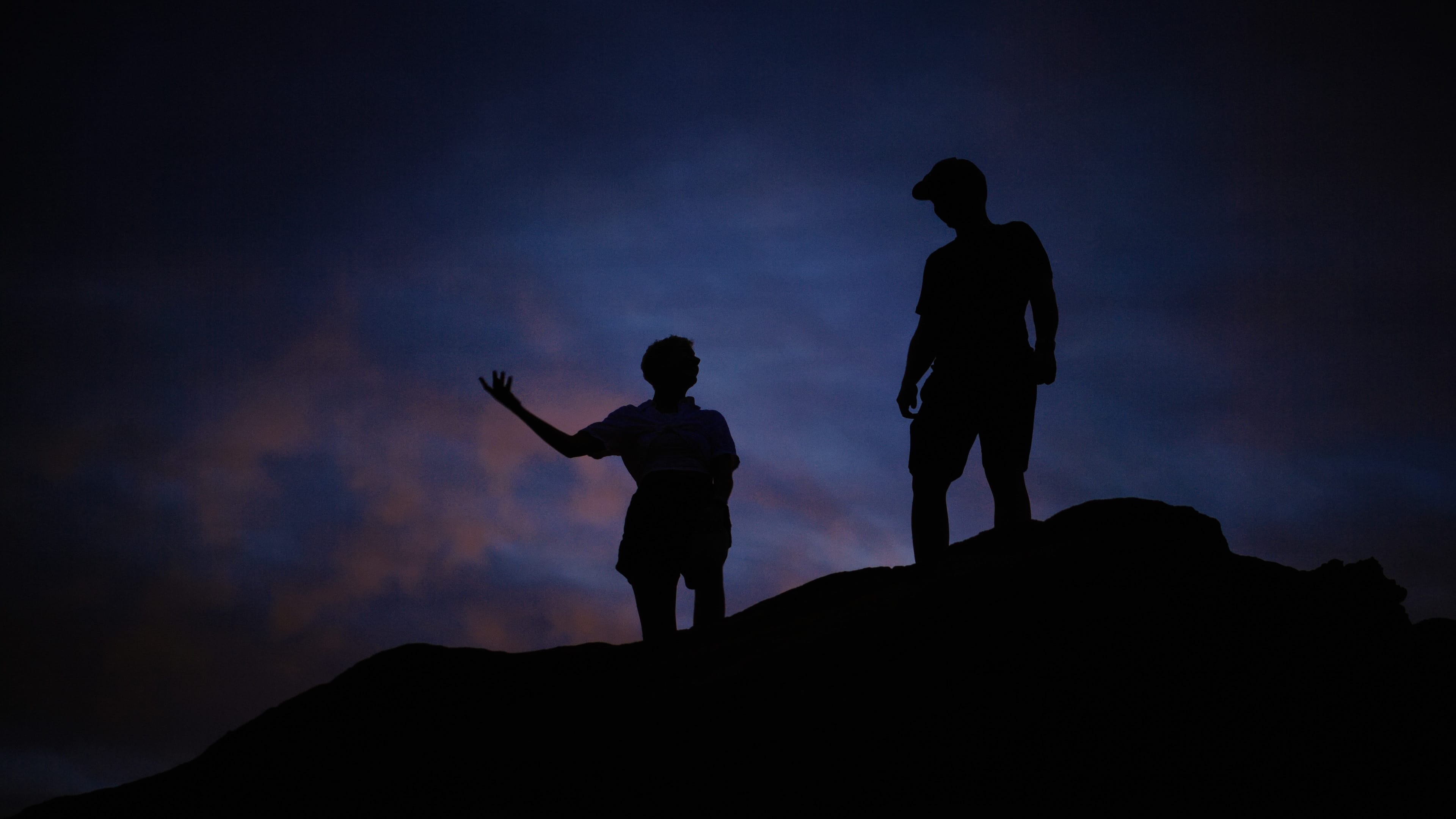 A film director sits in a chair facing a bustling, warmly lit night market scene. A camera setup is nearby, capturing the energetic atmosphere.
