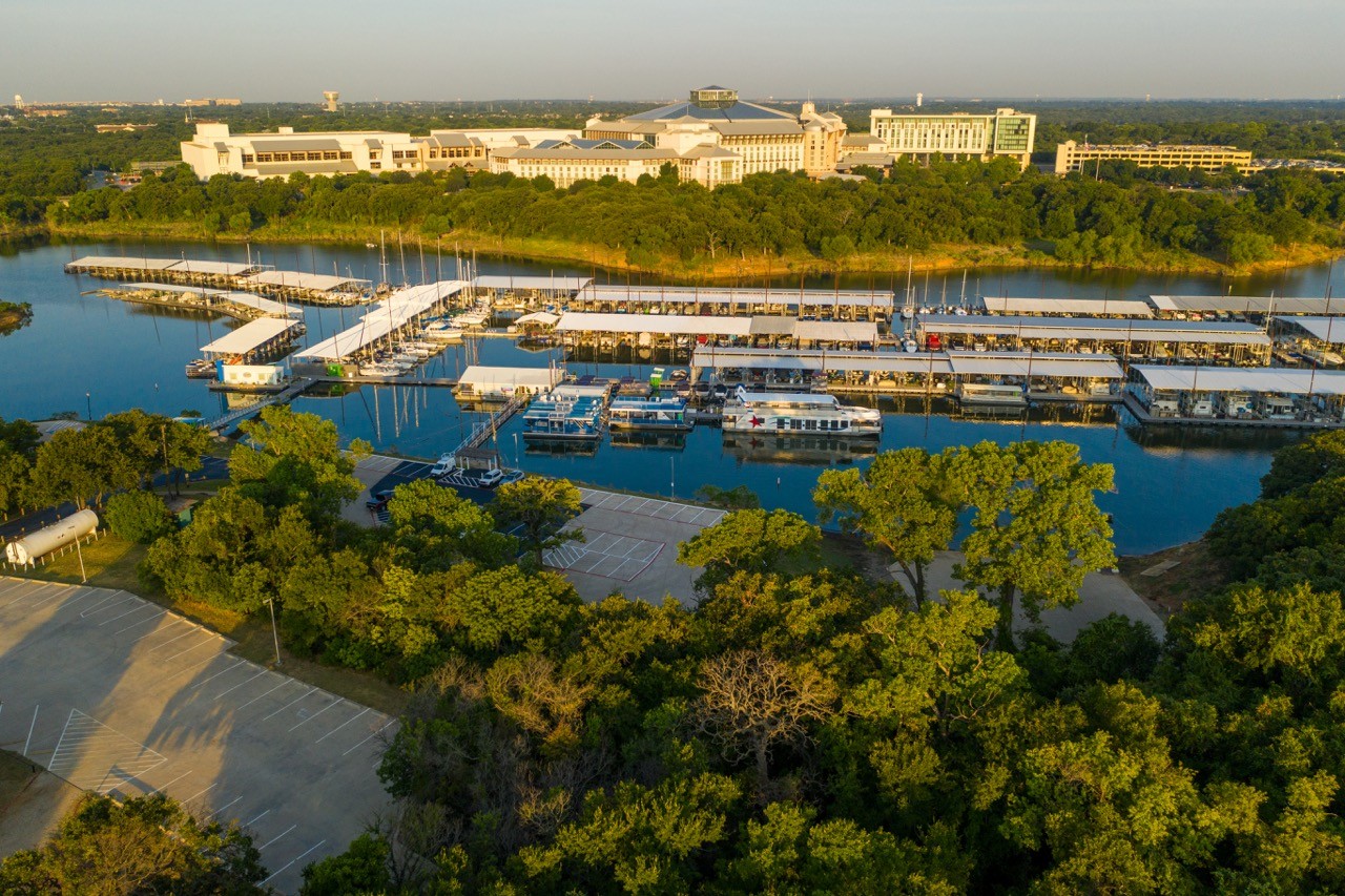 Aerial view of a marina with numerous docks and boats on a calm body of water, surrounded by lush greenery, with a large building complex in the distance, under a clear blue sky.