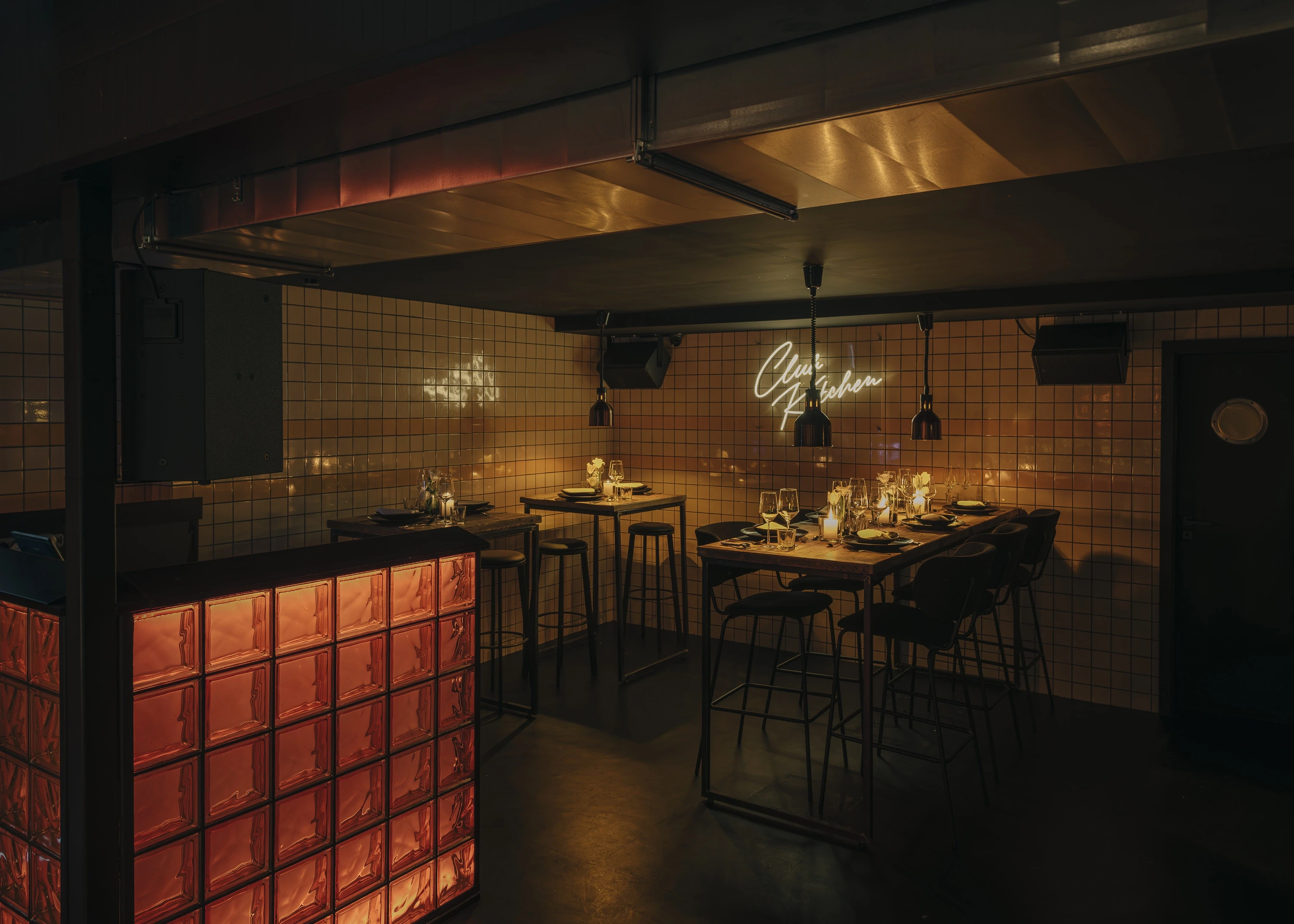 Club Kitchen at NATE CLUB with a glowing orange glass-brick counter, beige tiled walls, black high tables set for dinner and a Club Kitchen neon sign.