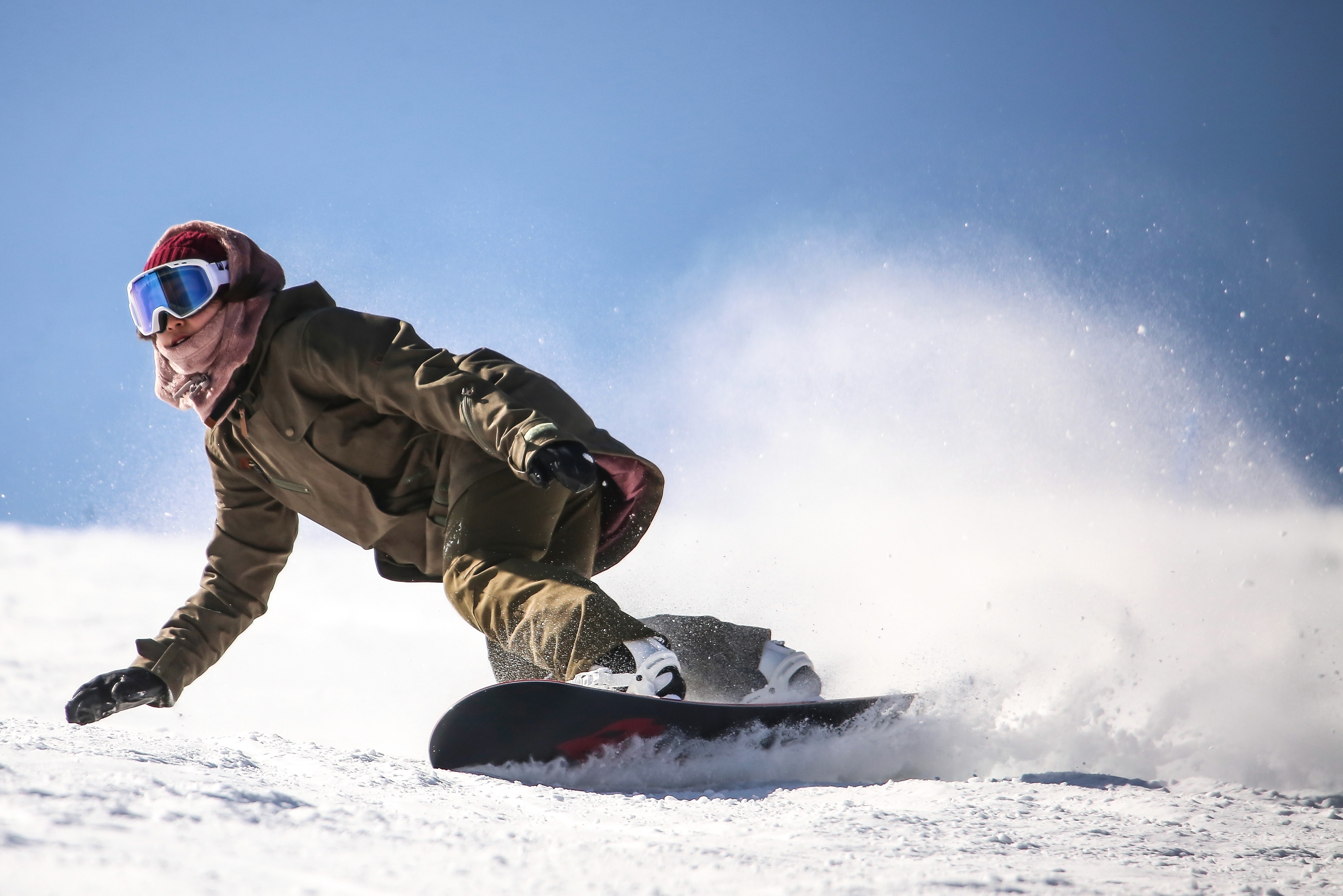A snowboarder carves through fresh snow, kicking up powder against a bright blue sky.