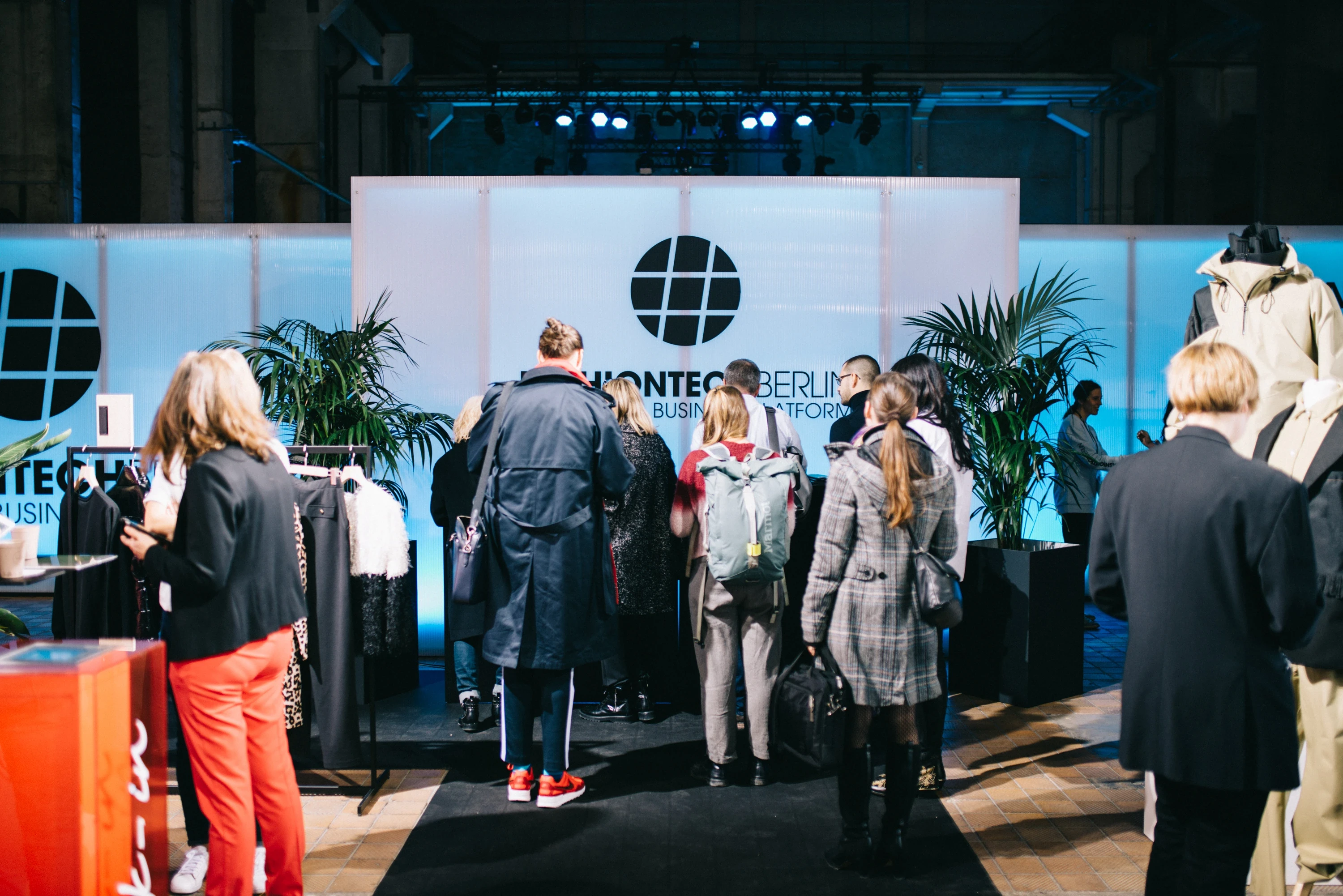 Visitors at the backlit FASHIONTECH Berlin logo wall between clothing racks and palm plants at the conference.