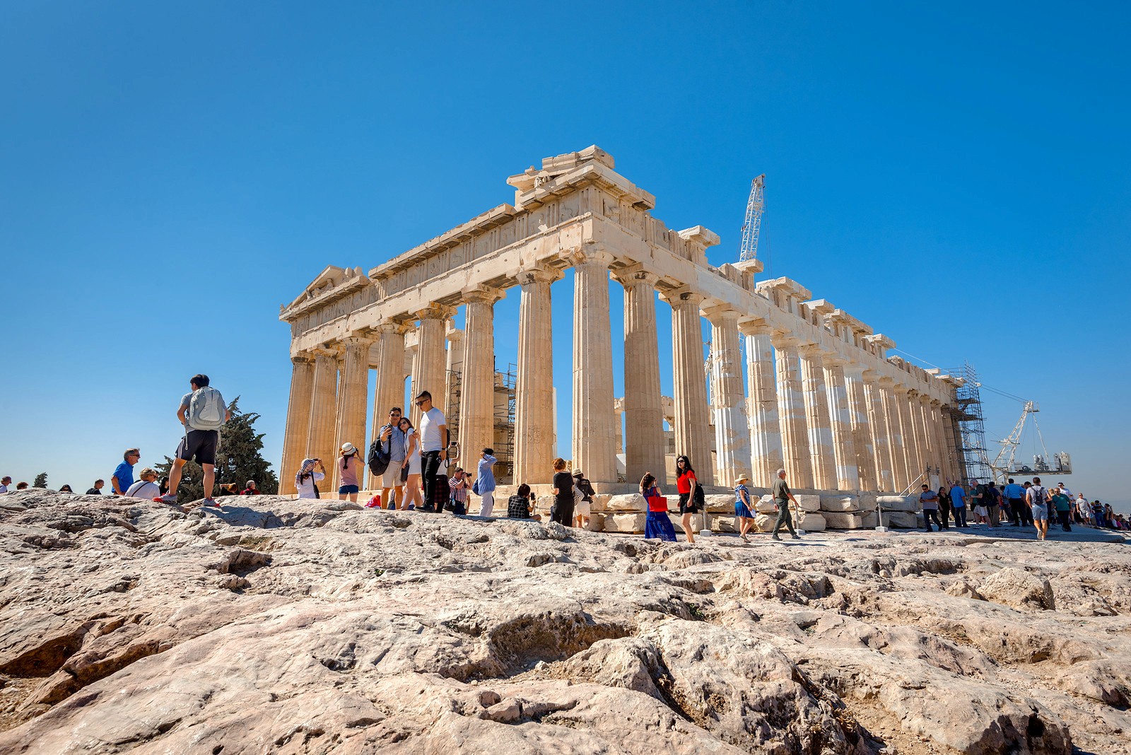 Crowds of tourists visit the ruined Parthenon temple on a rocky hill under a clear blue sky in Athens.