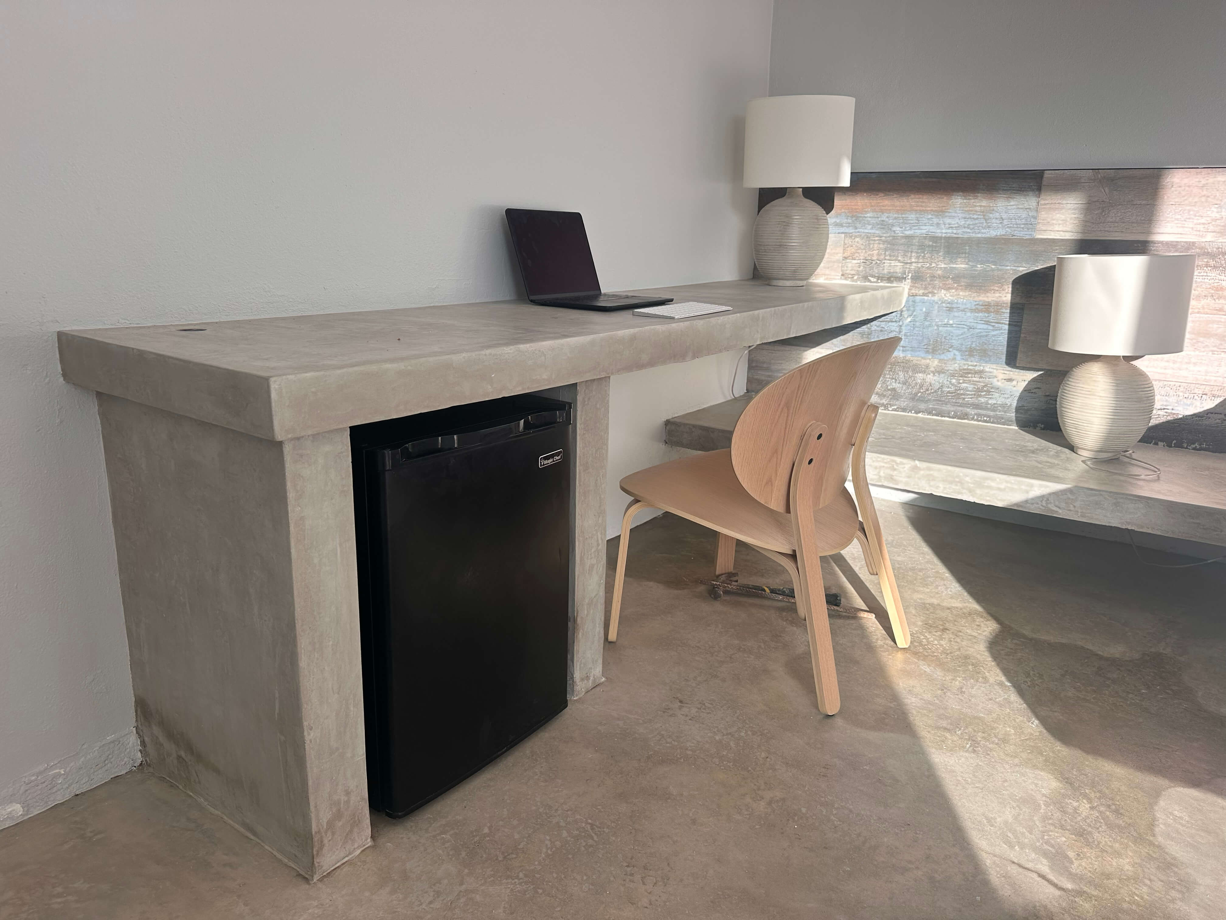 Room workspace with a concrete desk, mini-fridge, and a modern chair in Club Vieques room 1, featuring natural lighting.